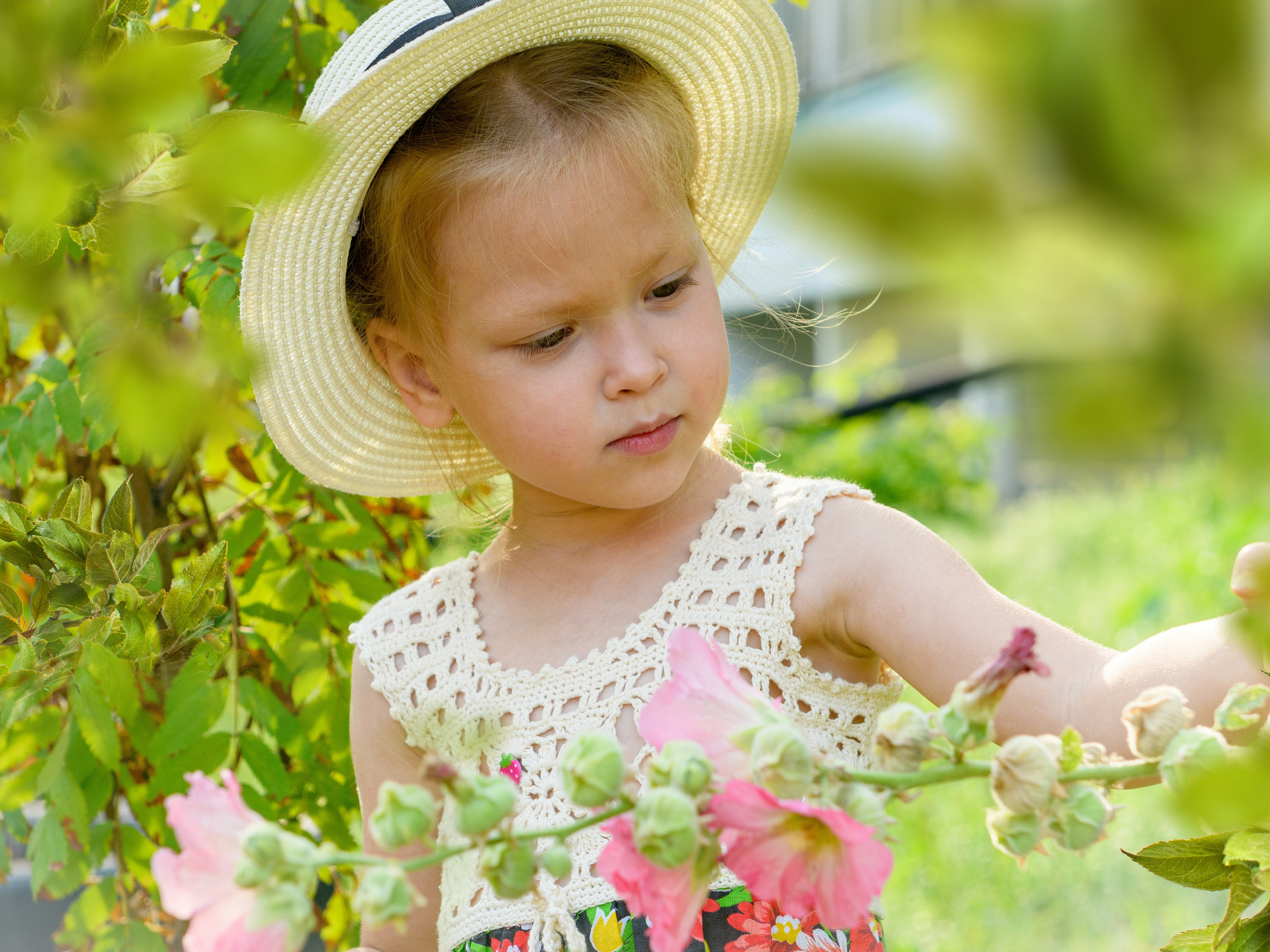 девочка в летней соломенной шляпке стоит и рассматривает цветок