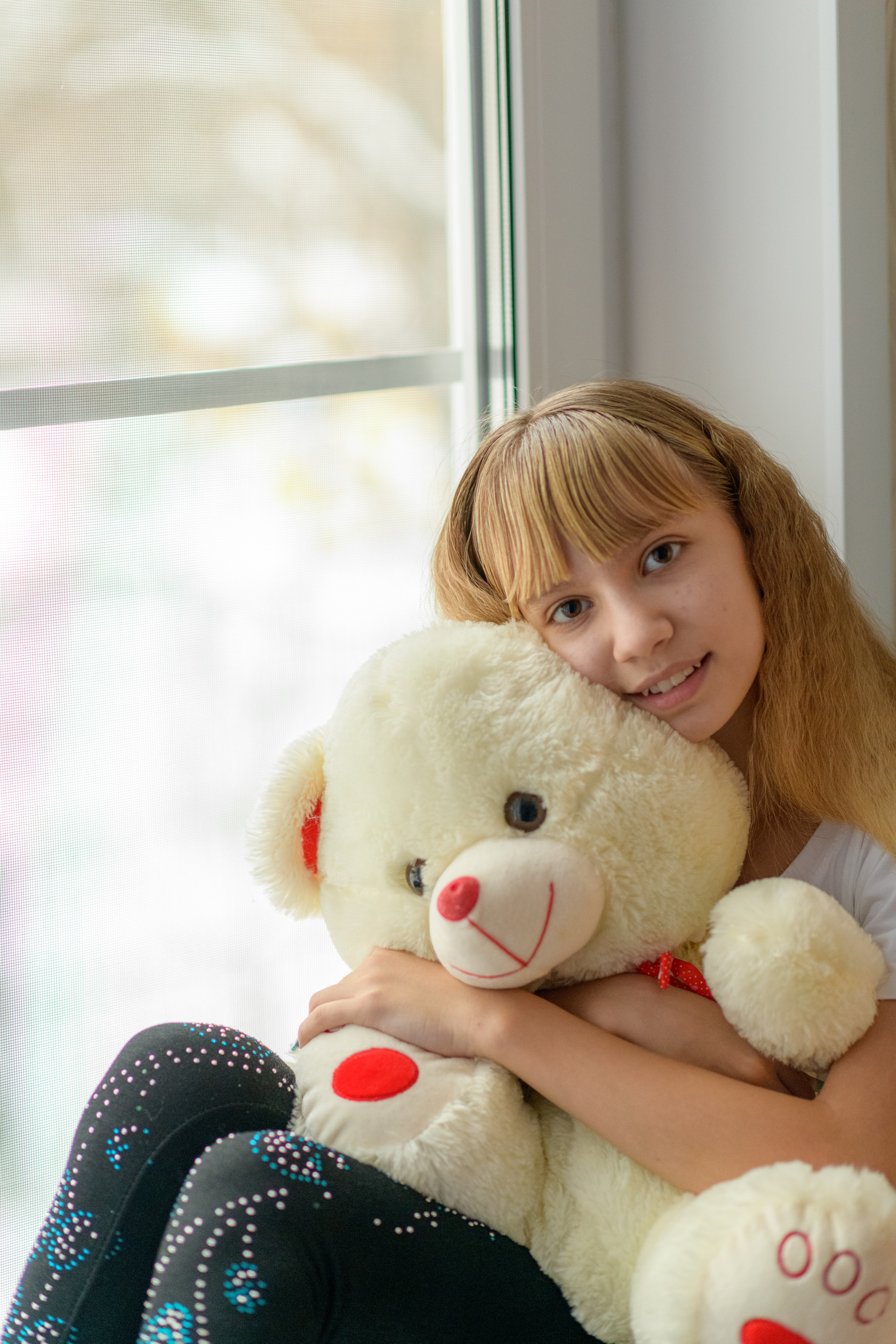 девочка со светлыми волосами сидит на подоконнике и обнимает большого плюшевого медведя