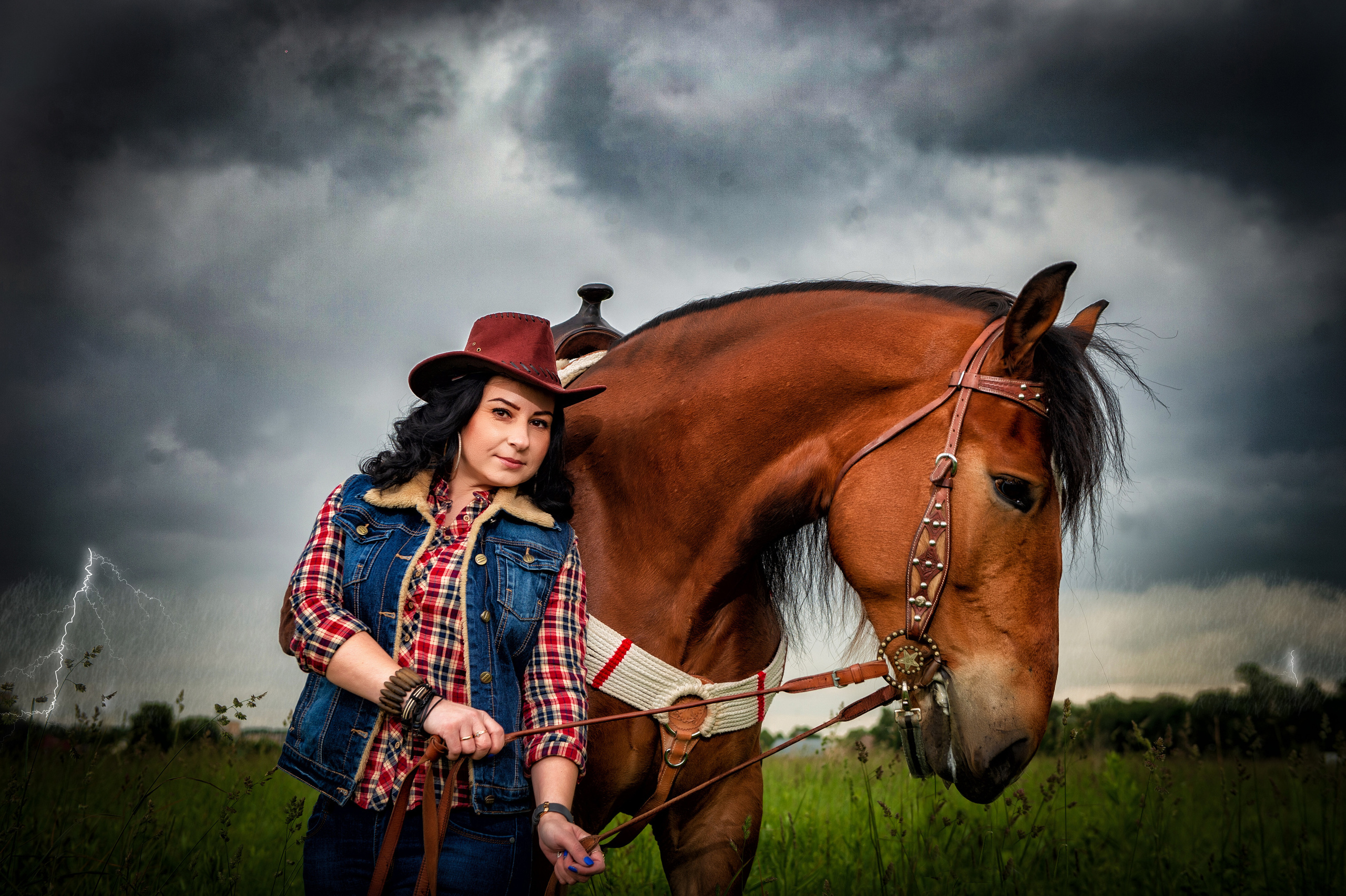 девушка в джинсовой жилетке и ковбойской шляпе стоит держа коня под уздцы рыжего коня
