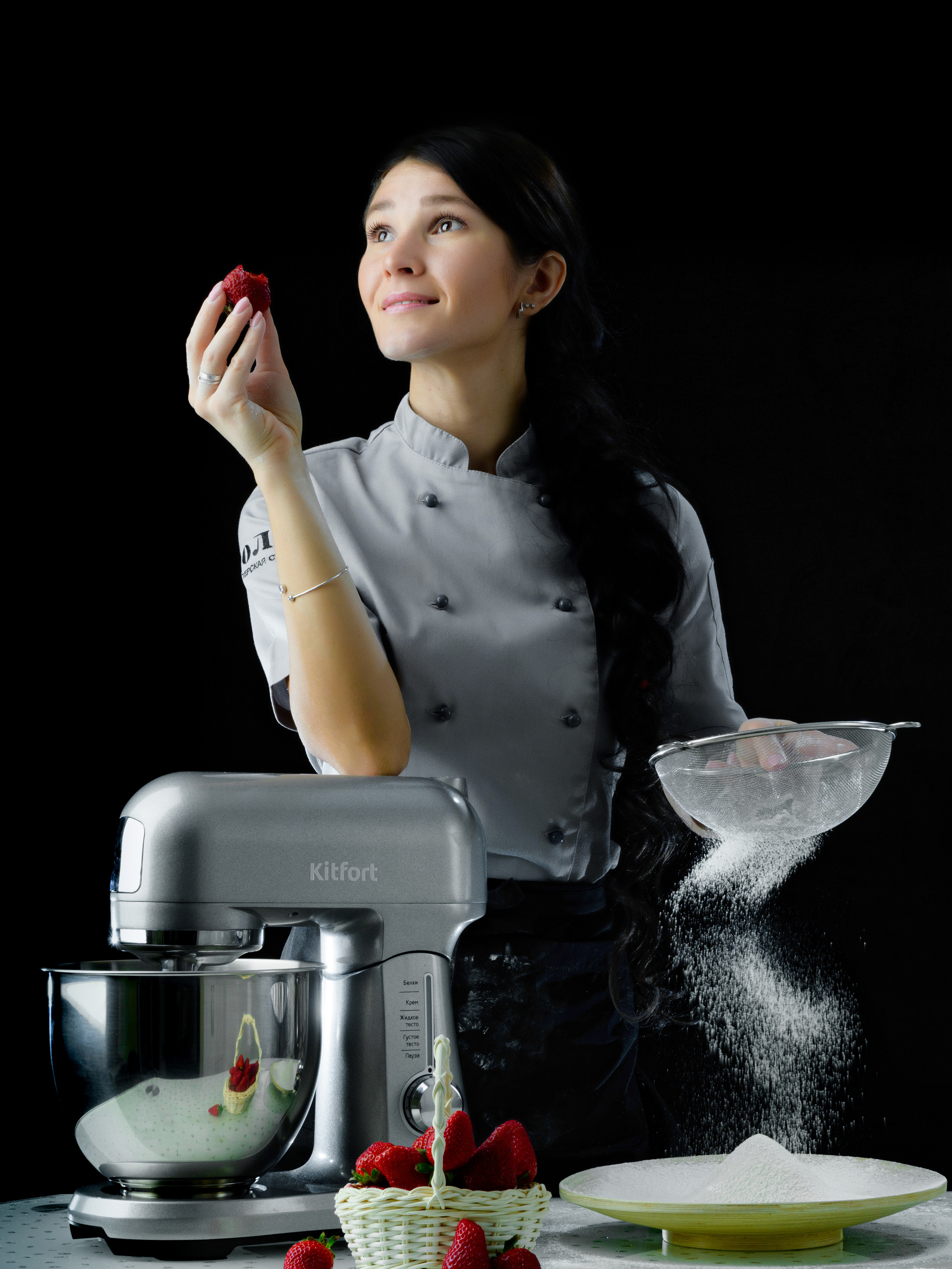 на черном фоне фото девушки просеивающей муку на столе стоит миксер и миска с клубникой