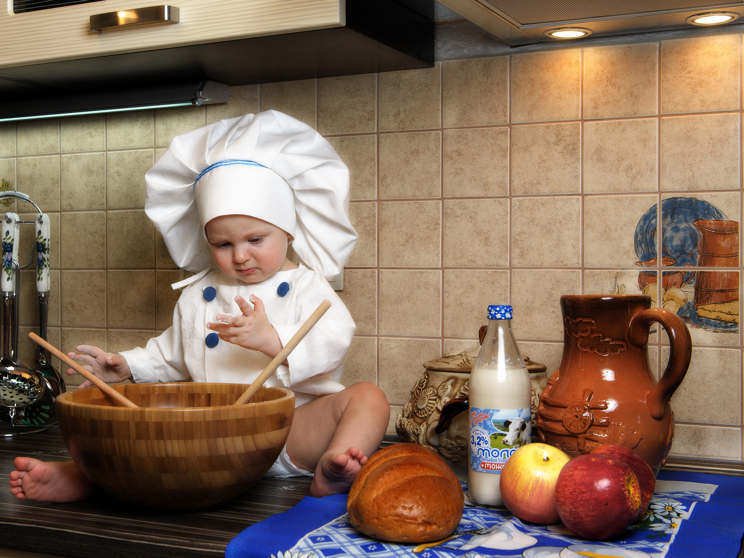 мальчик одетый в поварскую курточку и поварской колпак сидит на кухонном столе