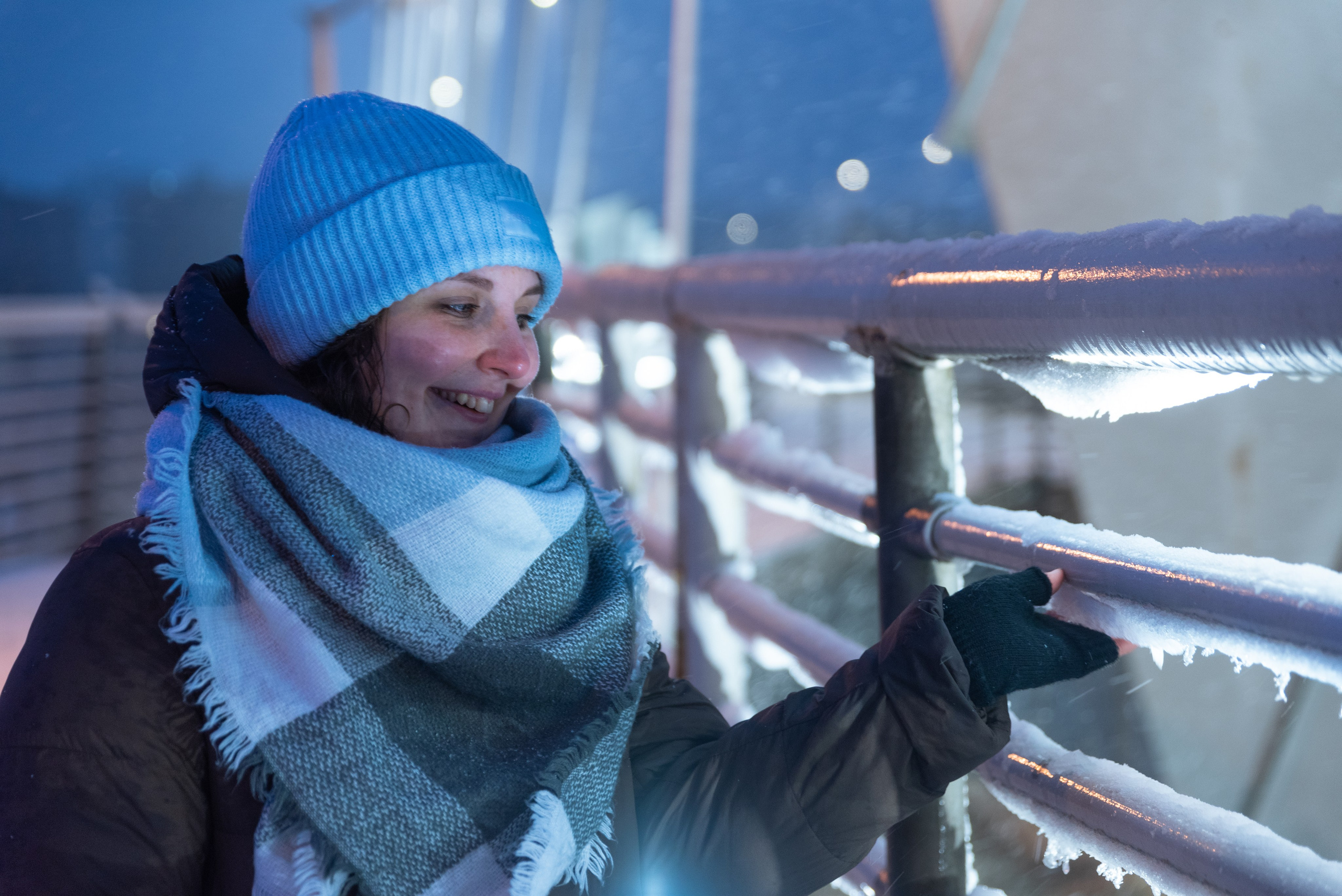 Ночь, зима, снег, свет фонарей. Девушка в широком шарфе и шапке стоит у обледеневшего металлического ограждения и отламывает кусочек льда.