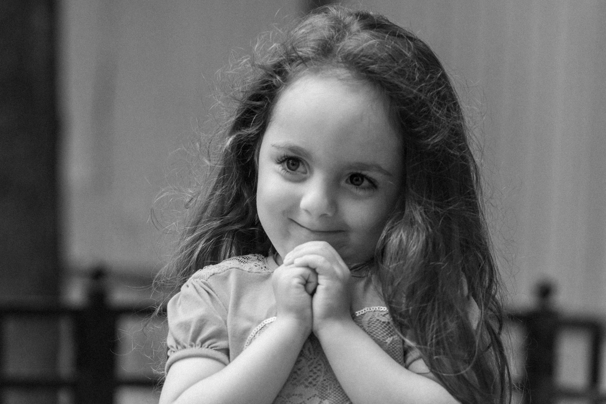 Черно-белый портрет маленькой девочки с длинными волосами. Она сложила руки под подбородком и застенчиво улыбается, смотря в сторону.