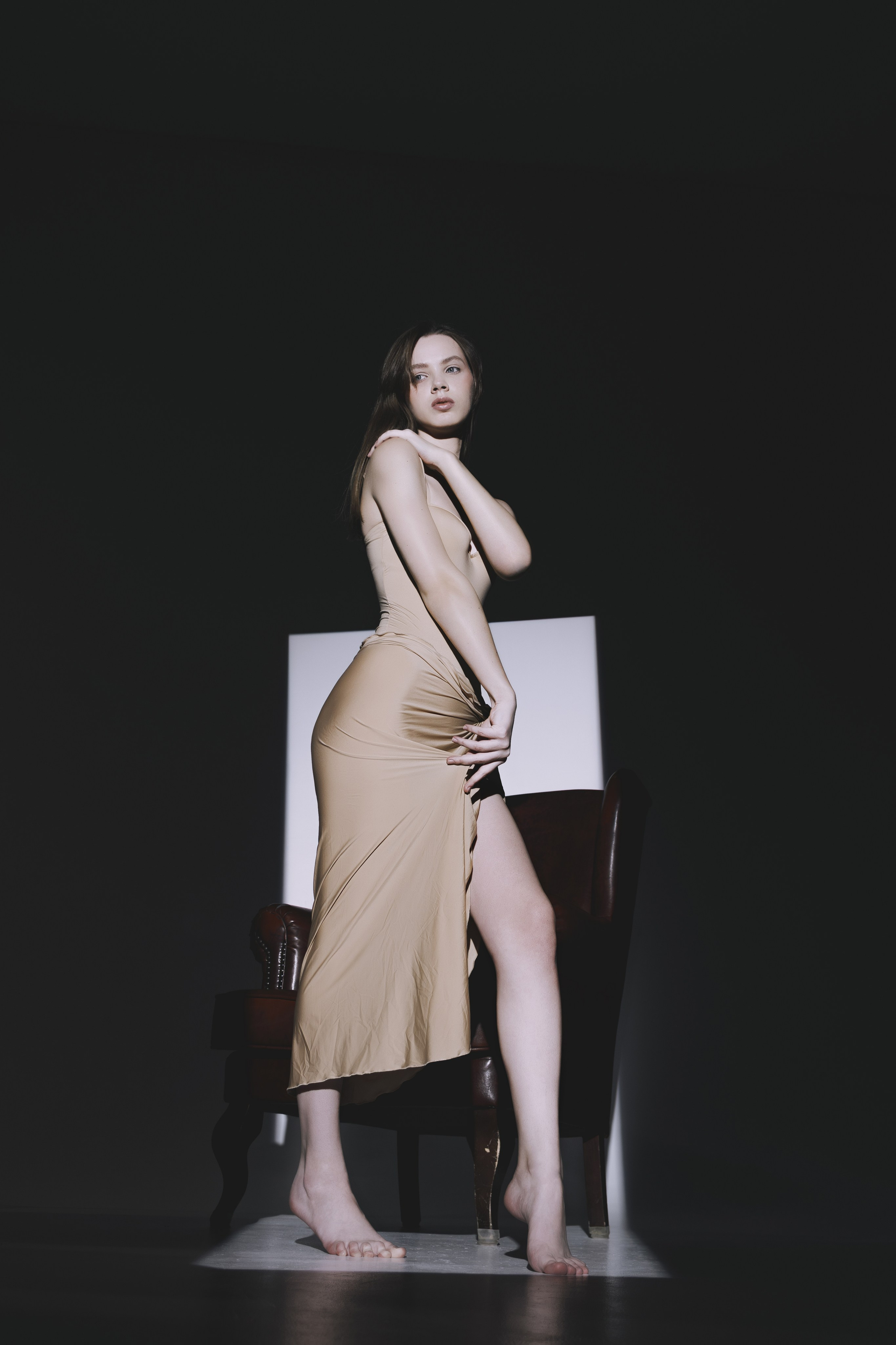 Художественная съёмка с акцентом на позу: девушка в бежевом платье позирует босиком, подчёркивая естественность и изящество. Классический стул становится частью композиции.