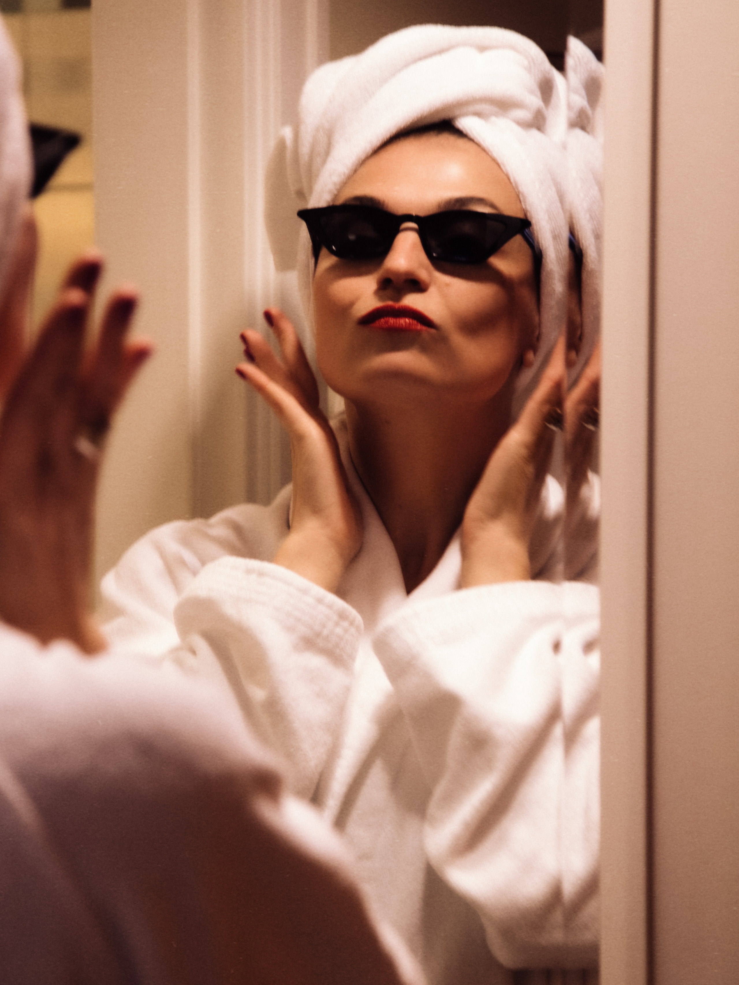 элегантная женщина в банном халате и тюрбане из полотенце в черных очках красуется  в зеркало