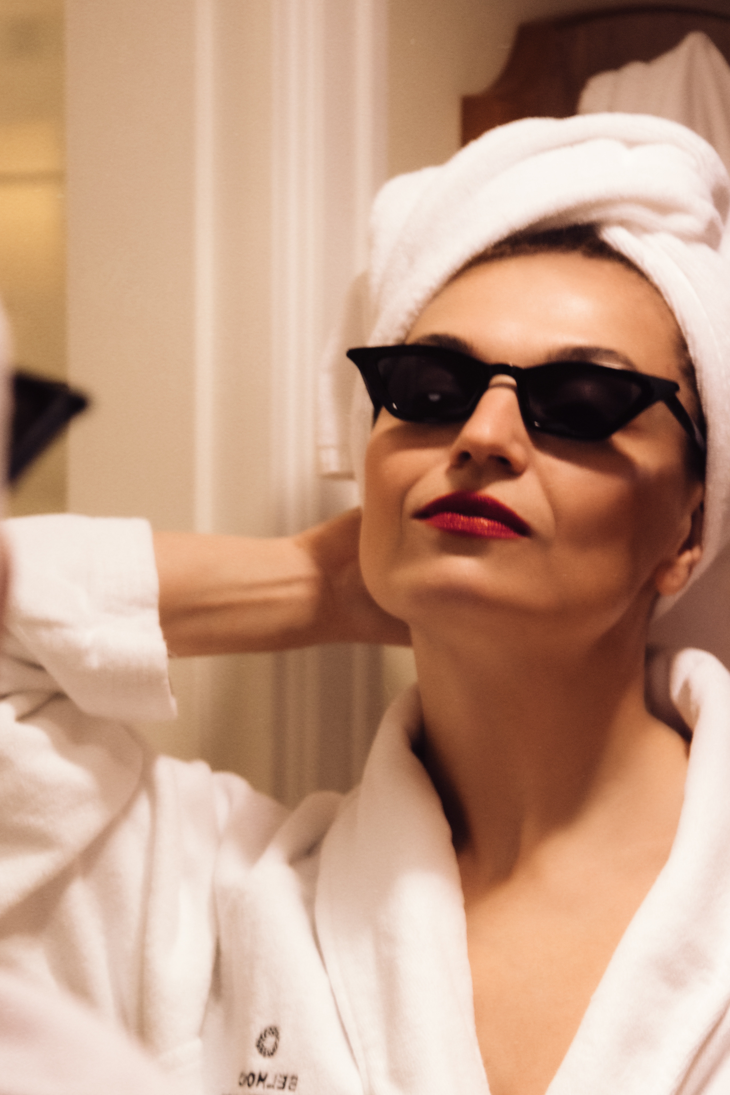 элегантная женщина в банном халате и тюрбане из полотенце в черных очках любуется на себя в зеркало