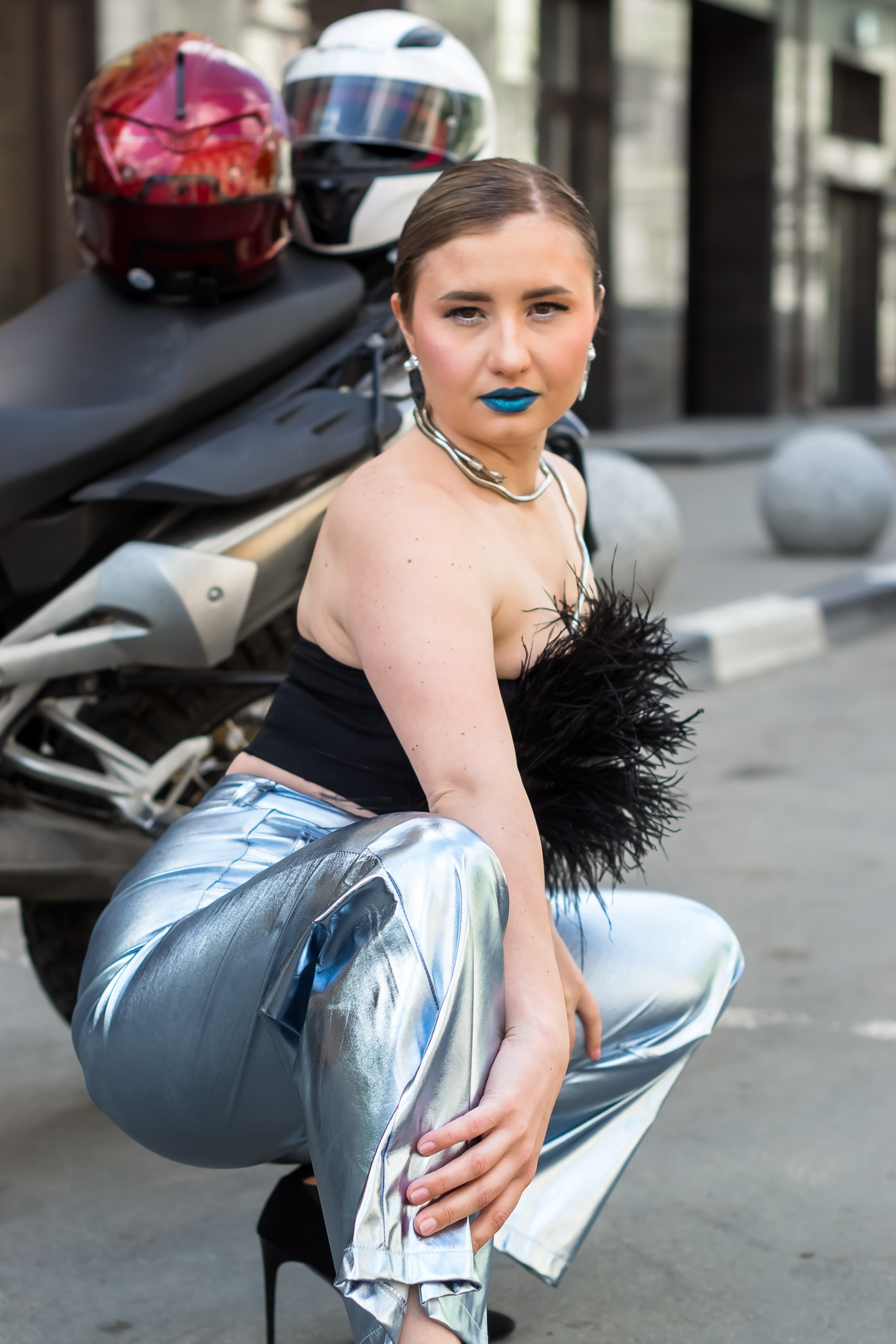 Портрет девушки в брюках и топике сидит на корточках около мотоцикла.