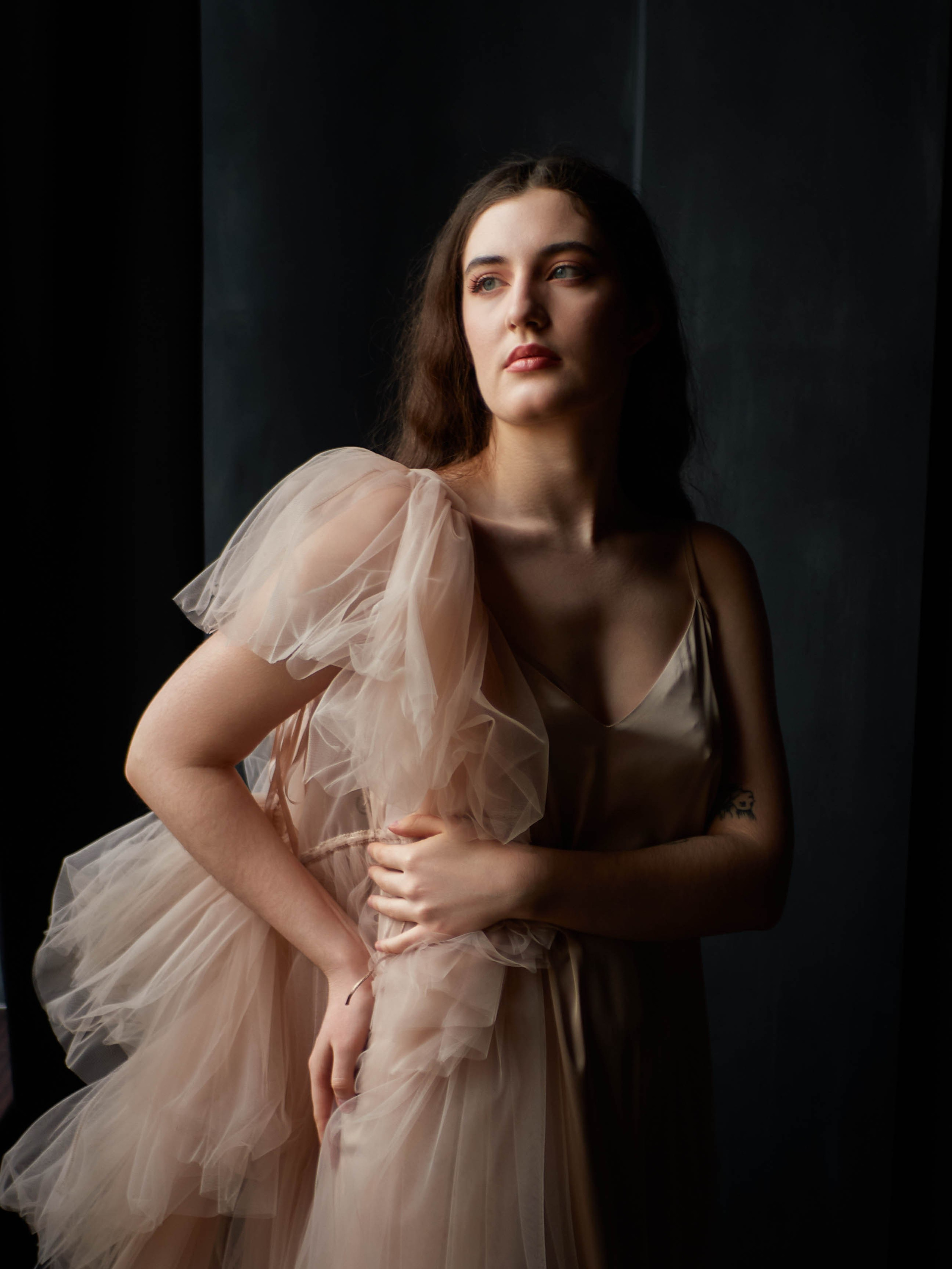 Девушка в бежевом платье на тёмном фоне с естественным светом.