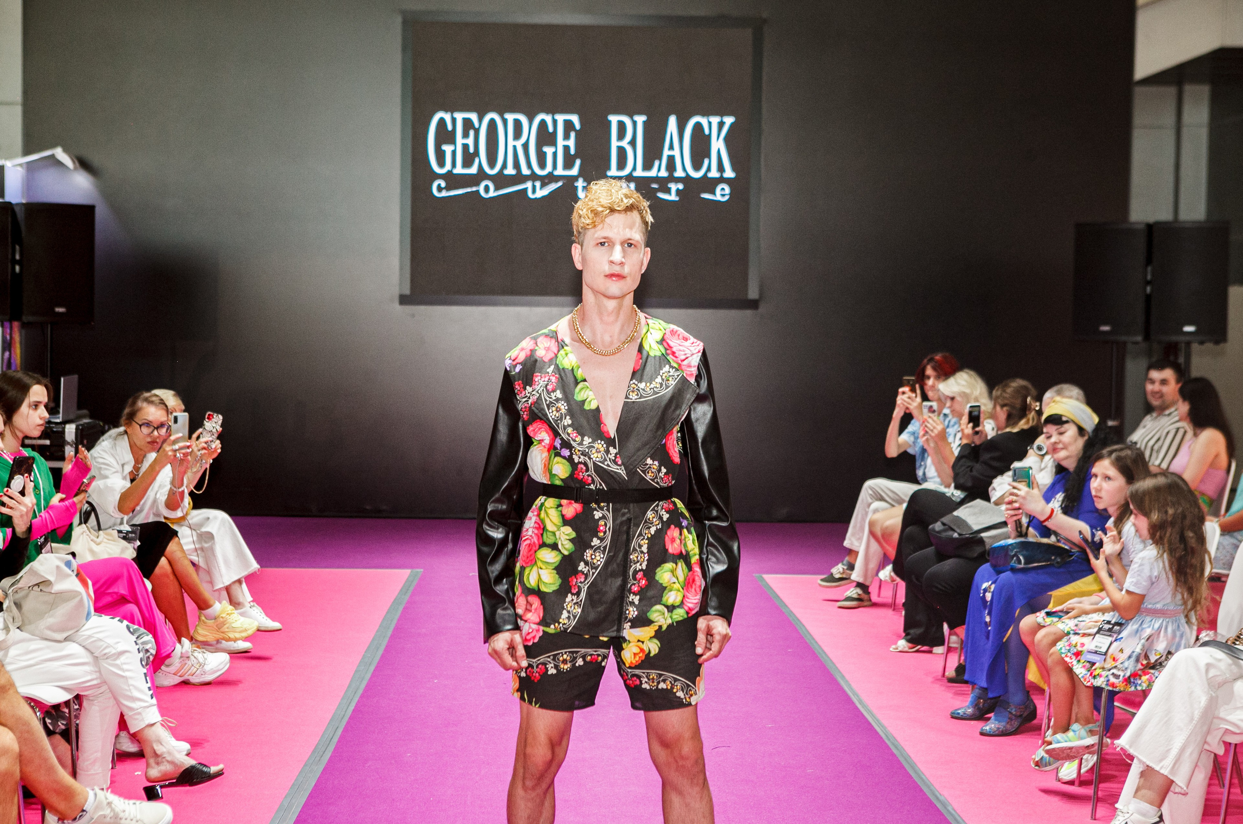  "Модели в ярких нарядах проходят по подиуму во время показа мод, освещенные яркими софитами и окруженные восторженными зрителями."