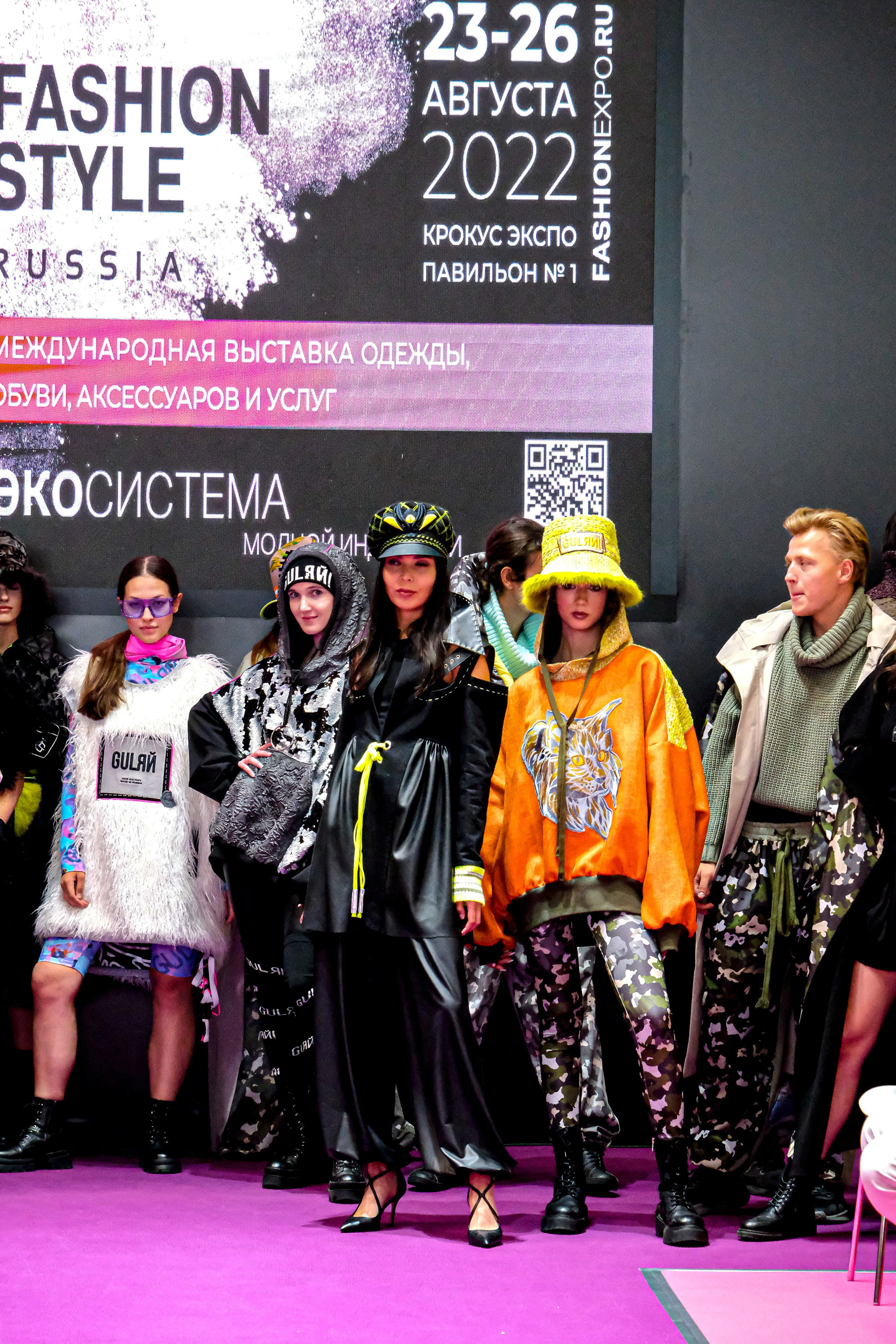  "Модели в ярких нарядах проходят по подиуму во время показа мод, освещенные яркими софитами и окруженные восторженными зрителями."