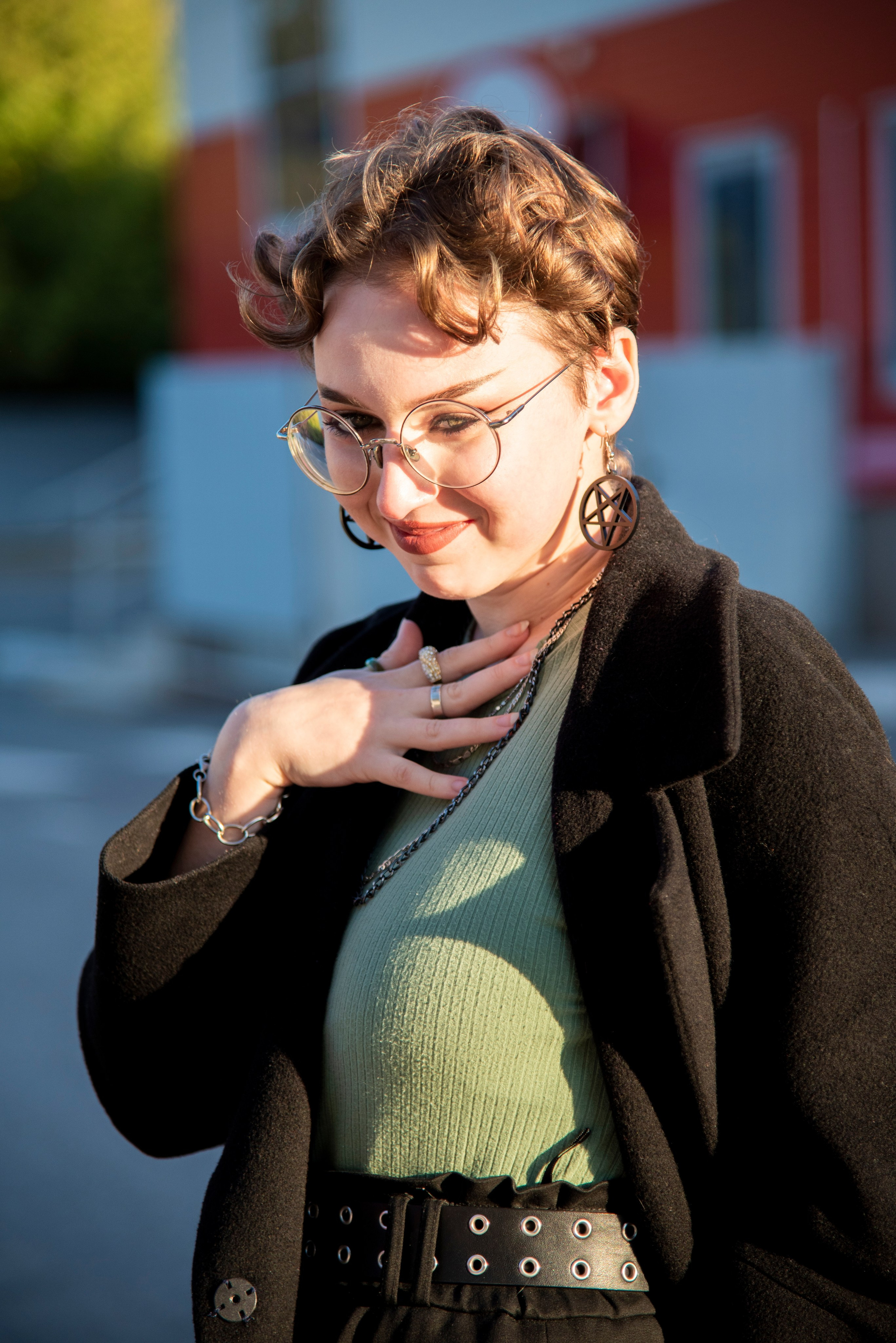 портет девушки. чёрное пальто. красивые позы. серьки в виде пентограммы. короткая стрижка. круглые очки. красный фон