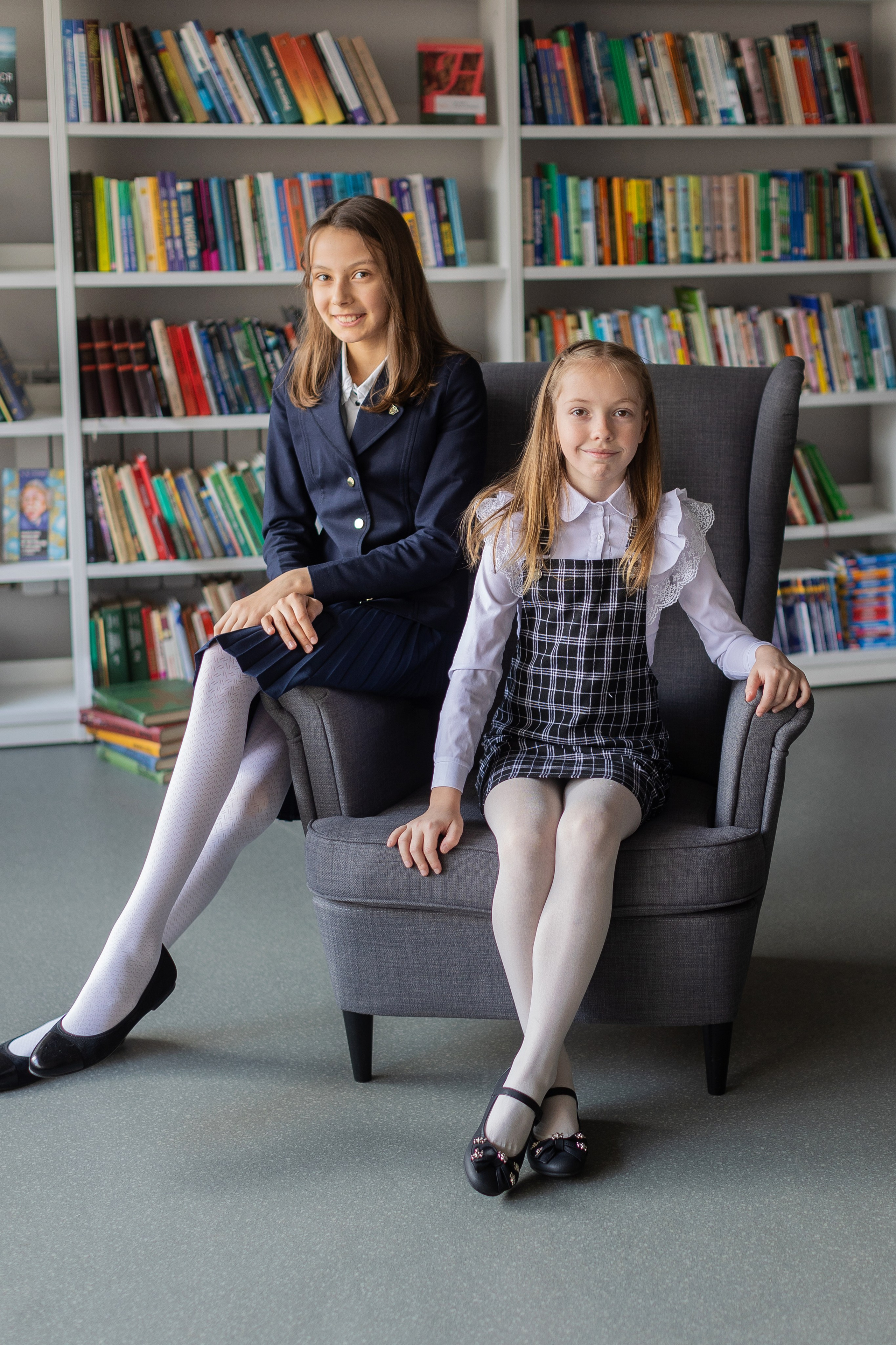 Две девочки сидят на стуле в библиотеке.