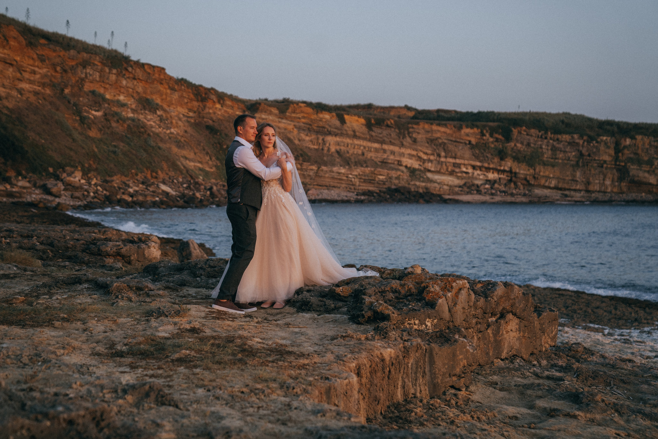 Счастливая пара жених и невеста целуется на фоне волн  скал и заката у моря океана.