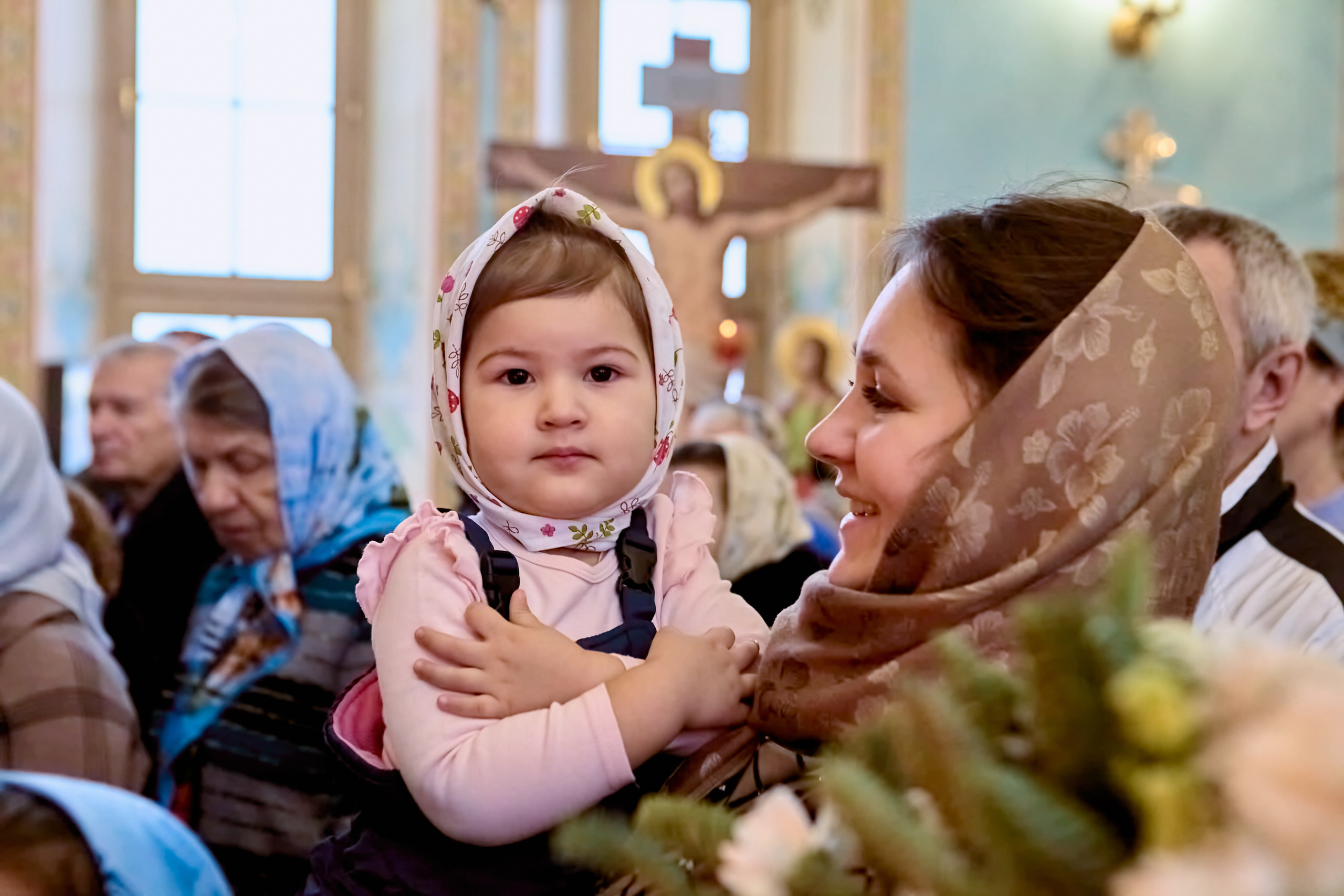Маленькая девочка на руках у мамы со сложенными крестом руками на груди в храме ждут причастия