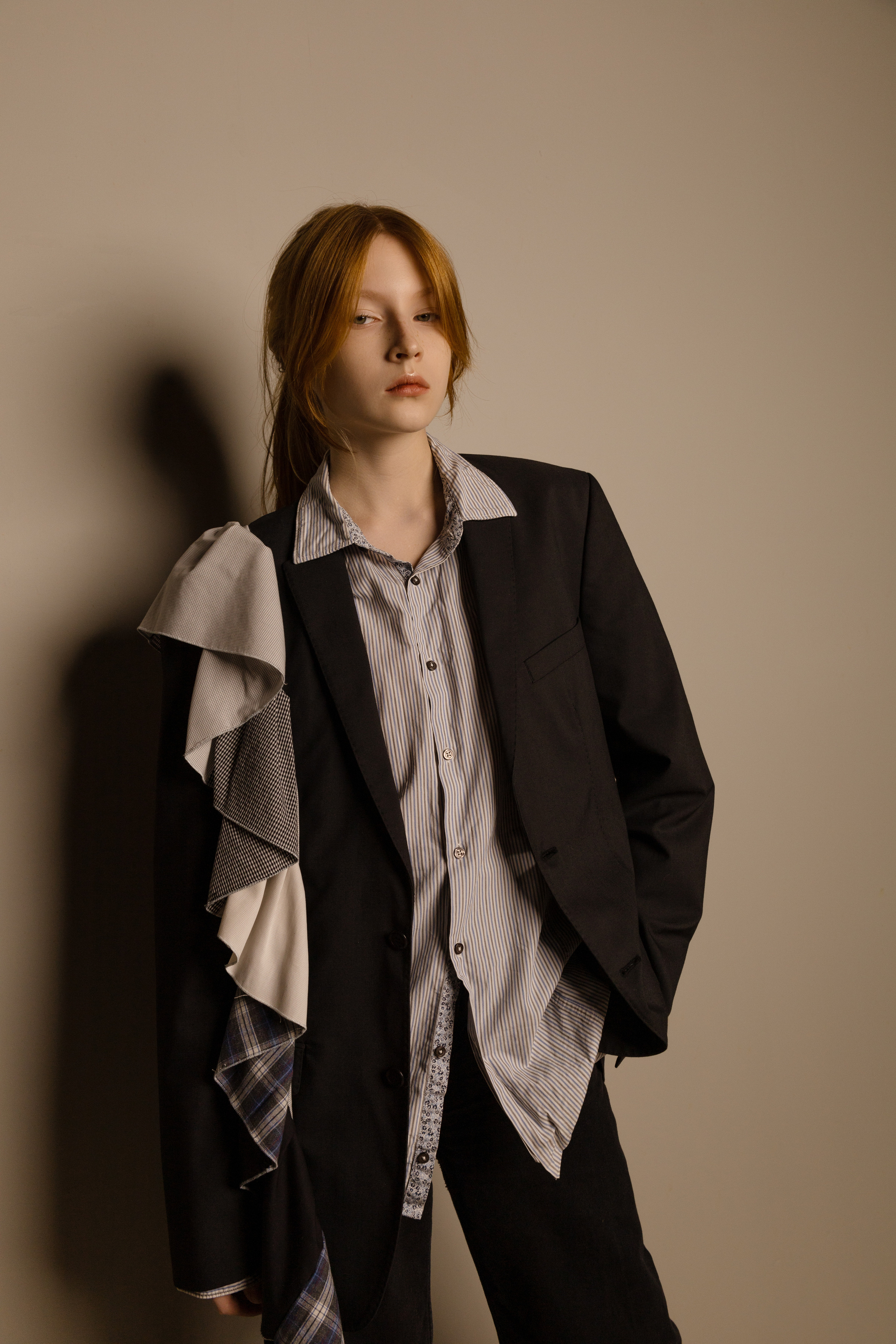 Девушка с  рыжими  волосами до плеч уверенно позирует на простом фоне. На нем черный блейзер, рубашка в полоску, черные брюки и светло-голубые туфли на каблуках.