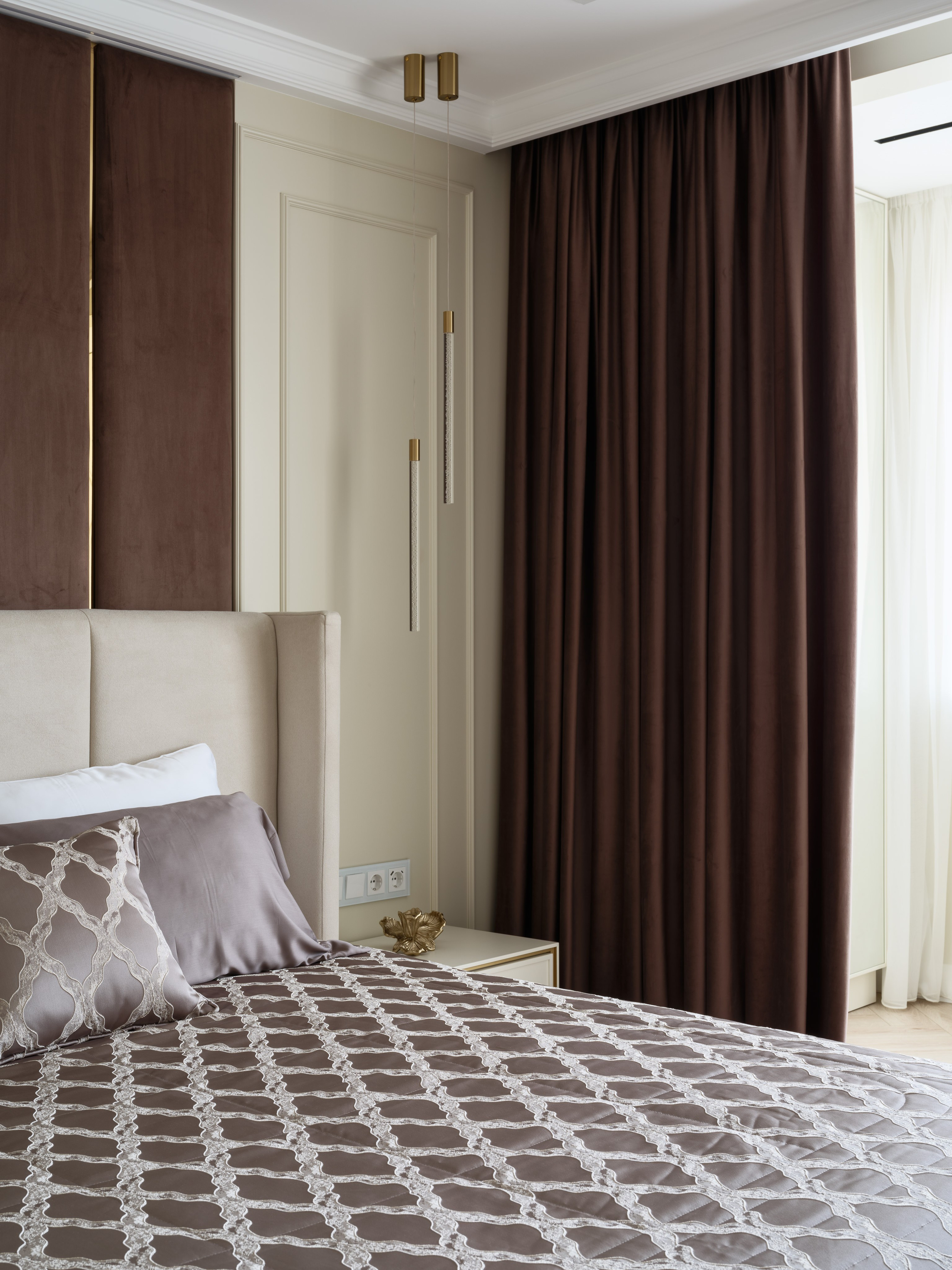 Современный интерьер спальни в спокойных тонах с высоким мягким изголовьем кровати, коричневыми бархатными шторами и подвесными светильниками с золотыми акцентами. Фото демонстрирует стильный и уютный дизайн, созданный Евгенией Ермошиной и снятый Сергеем Денисенко.