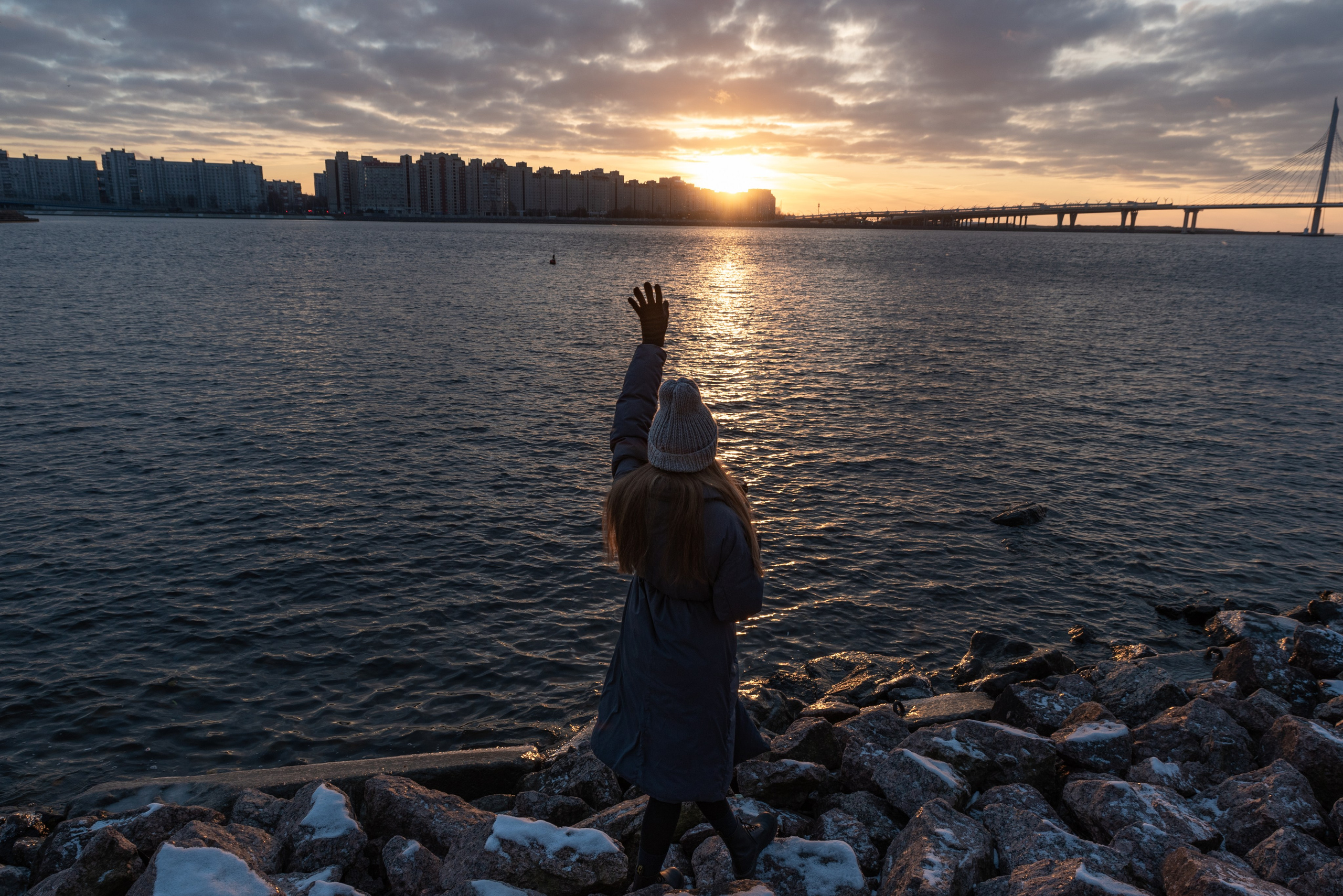 Берег, большие камни. Девушка стоит на камнях, протягивает руку к заходящему солнцу.