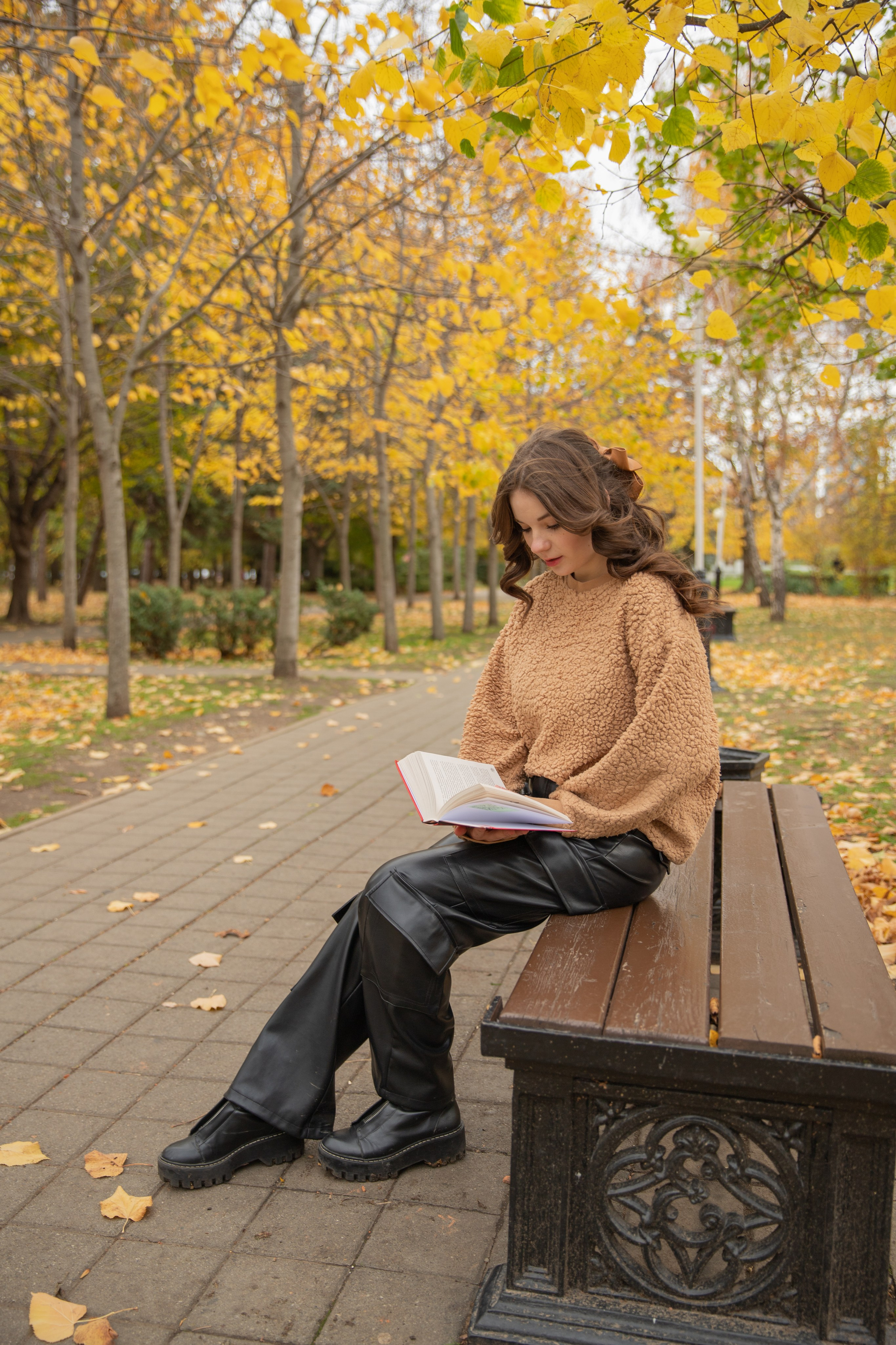 Красавица в уютном свитере  читает книгу на скамейке в осеннем парке Чистяковская роща, окружённая желтыми листьями и теплым светом.