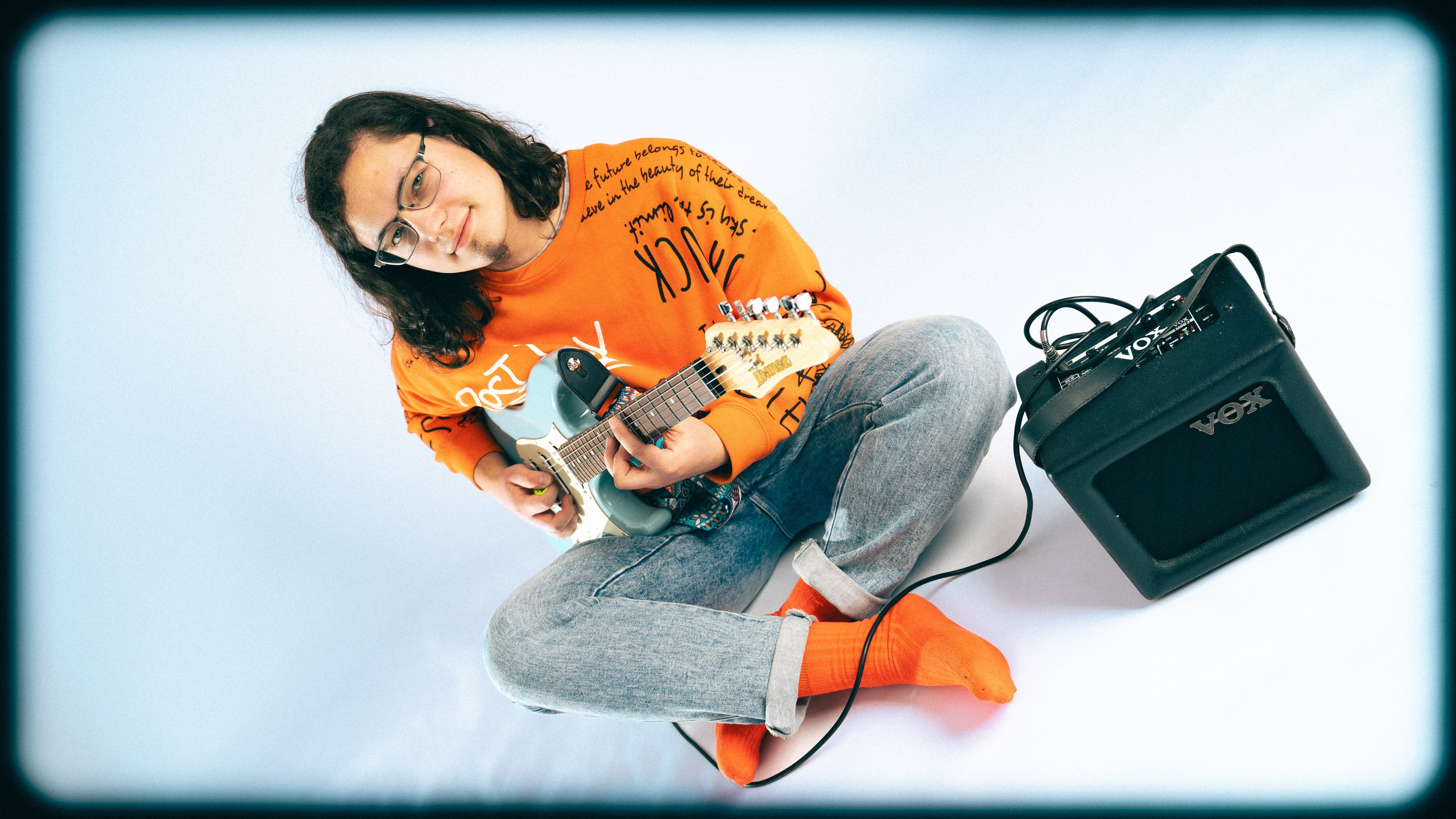 Молодой парень позирует в студии в яркой оранжевой одежде с гитарой