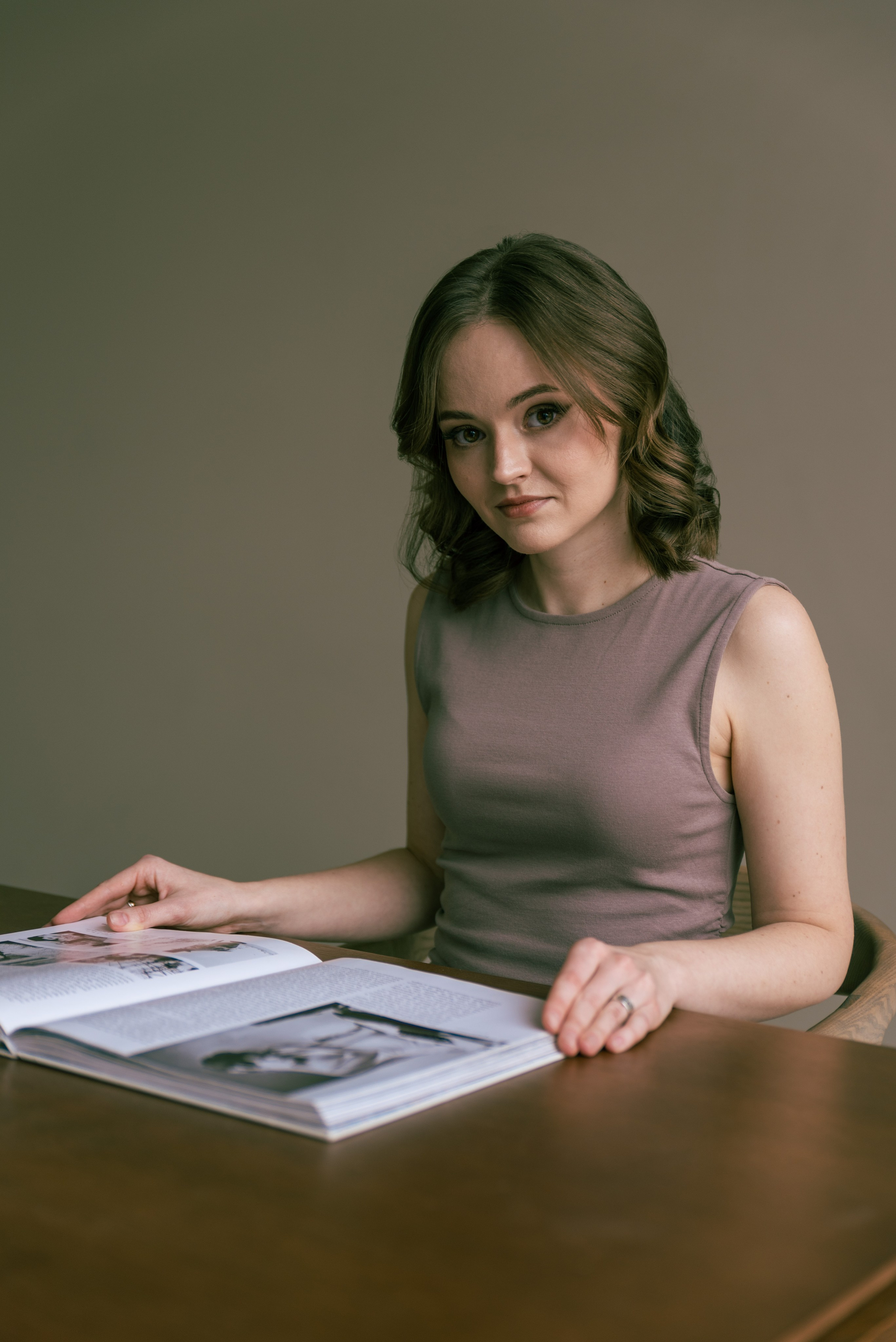 Девушка сидит за столом. Касается пальцами страниц большой книги. Смотрит на фотографа.