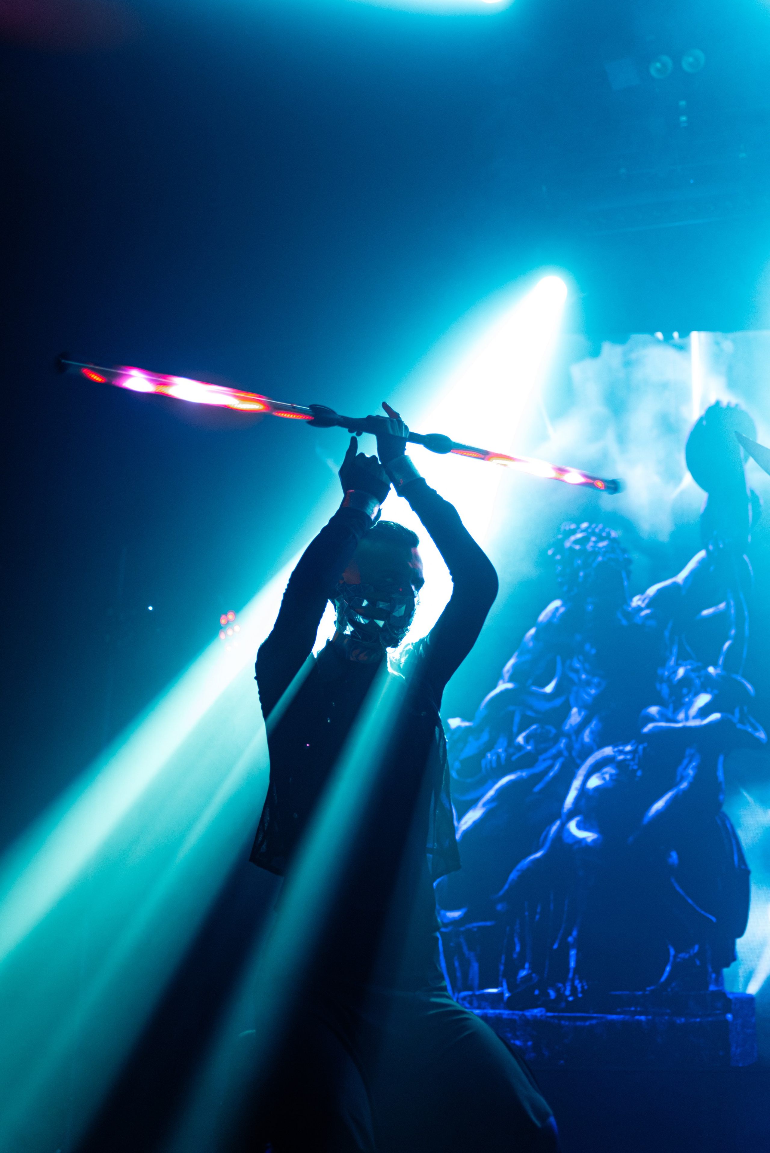 Танцор на сцене, чёрная одежда, на лице зеркальная маска в руках над головой длинная световая палка, на заднем плане изображение скульптуры в синих тонах, на человека сзади светит синий прожектор