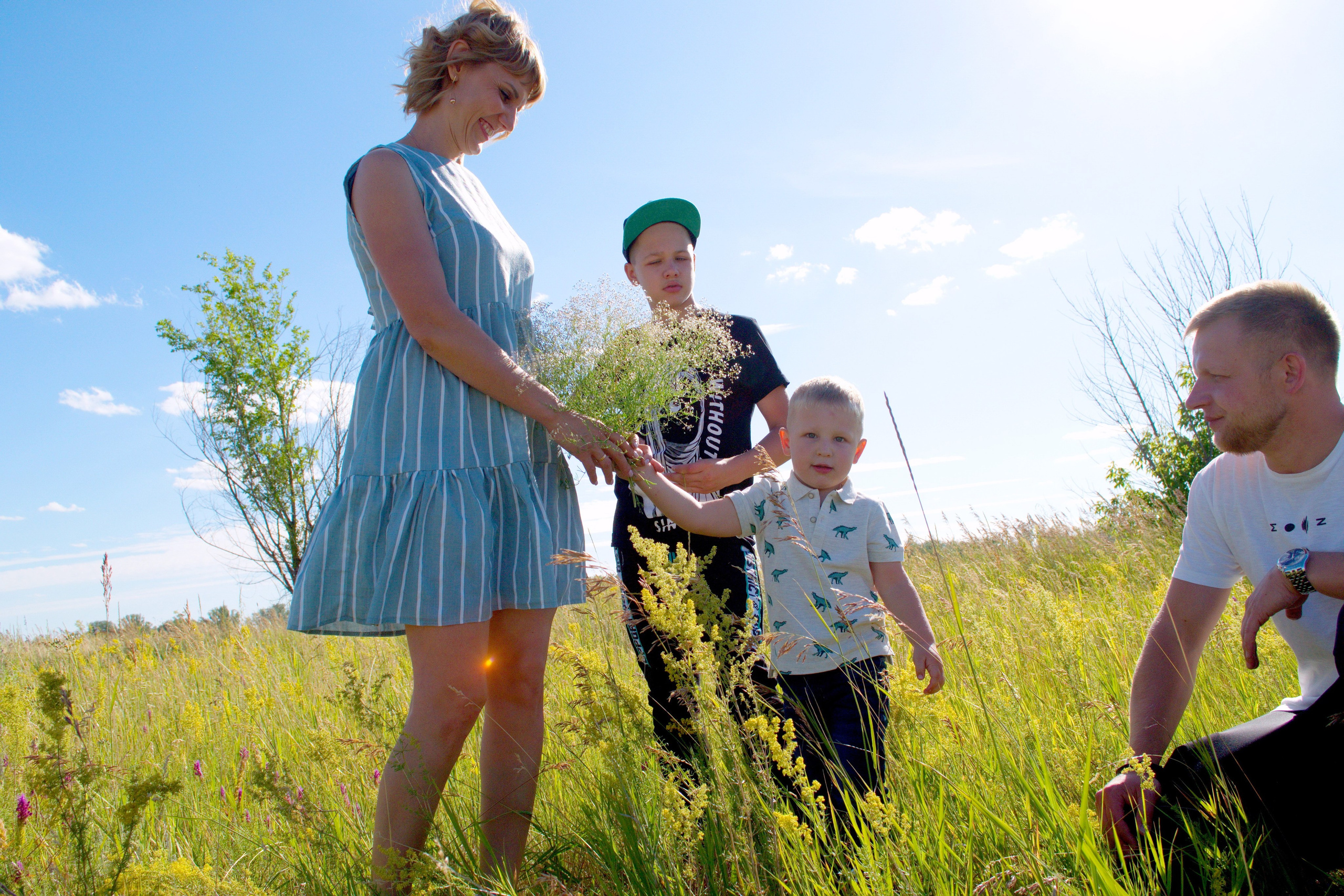 Маленький ребёнок дарит маме цветы на лугу. Отец и старший сын смотрят на них.