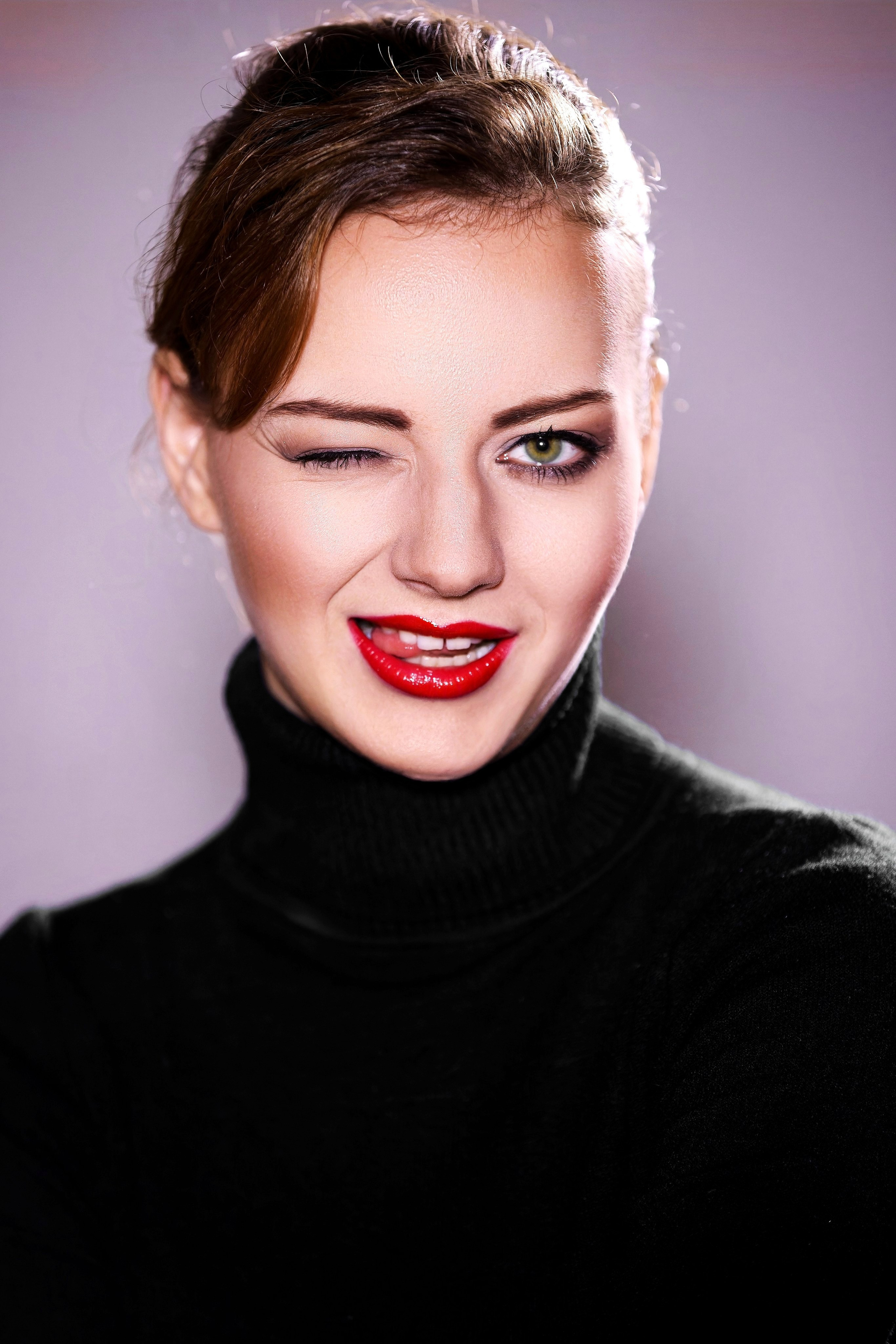 Крупный план портрета девушки с красной помадой и подмигиванием, черная водолазка, студийное освещение, серый фон, яркий макияж