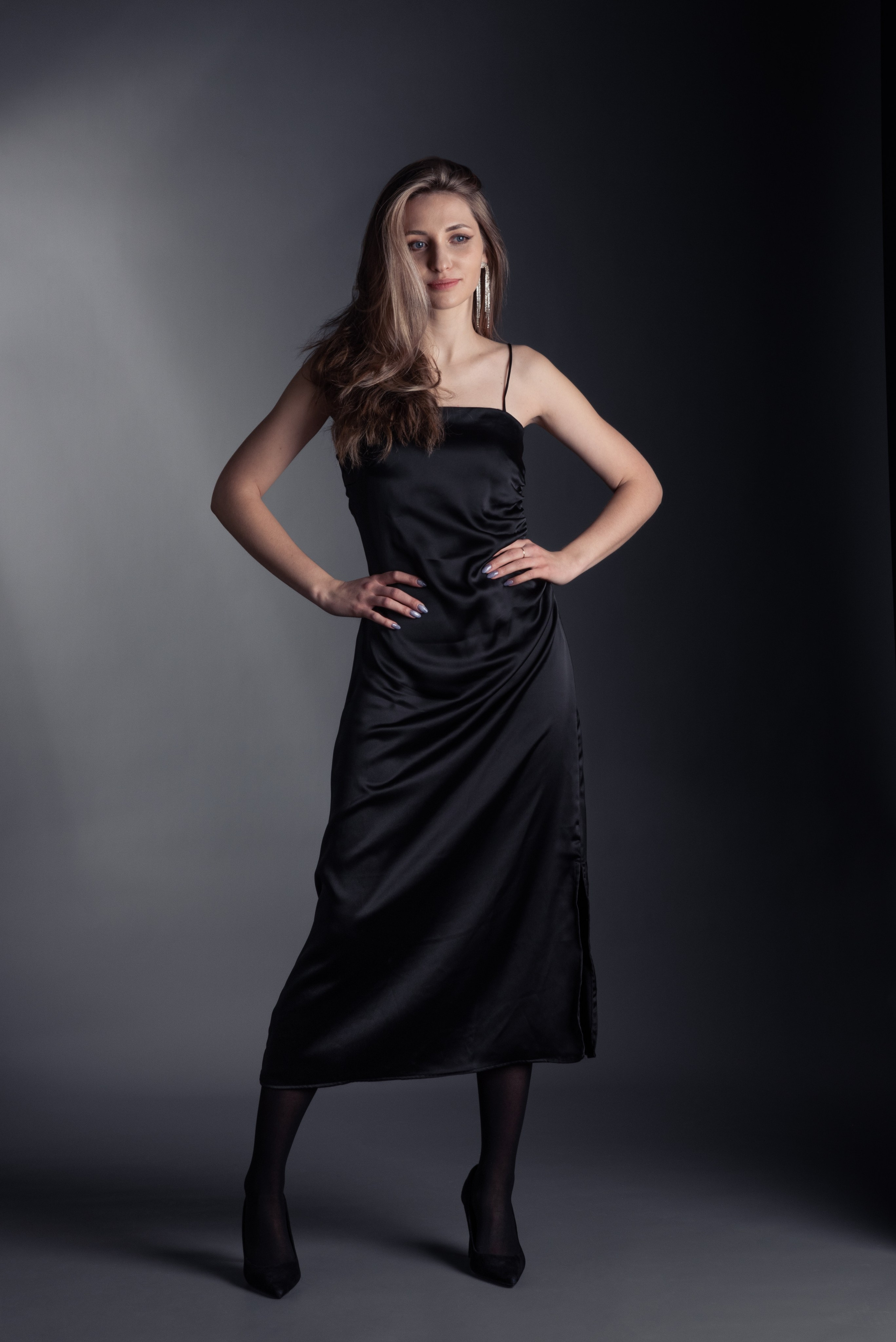Девушка в чёрном платье стоит в фотостудии, за спиной чёрный фон. Уверенная поза упирает руки в бока.