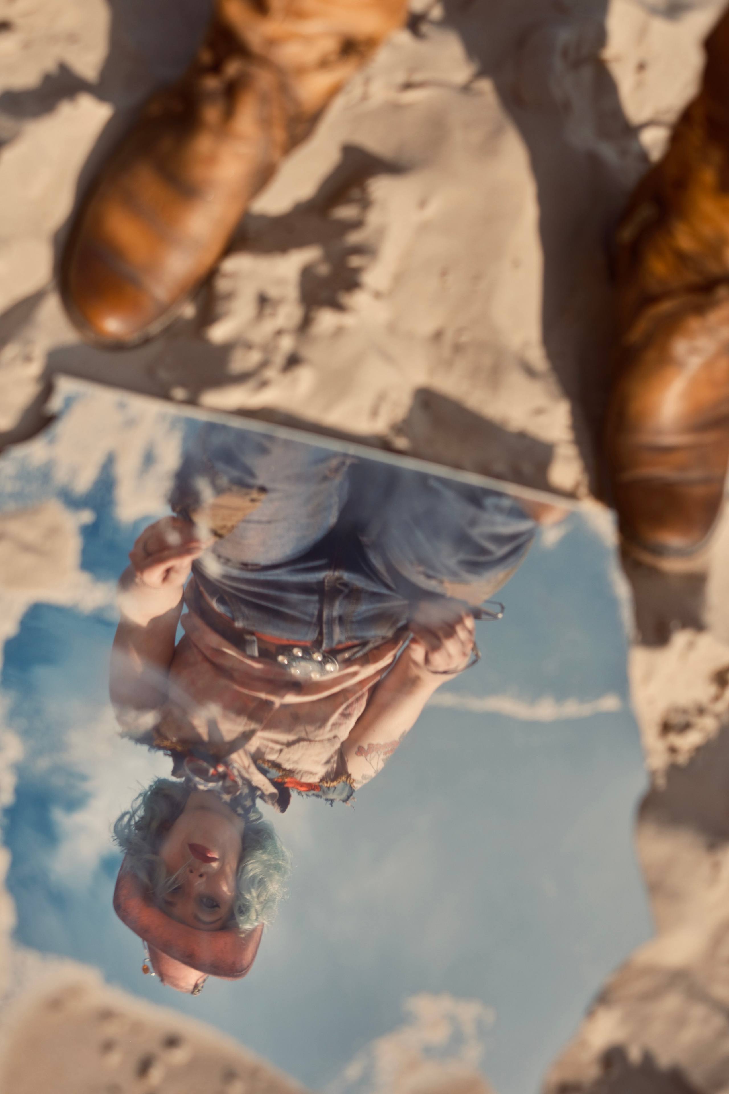 Девушка в розовой ковбойской шляпе смотрит в зеркало которое лежит на песке в стиле пост апокалипсис