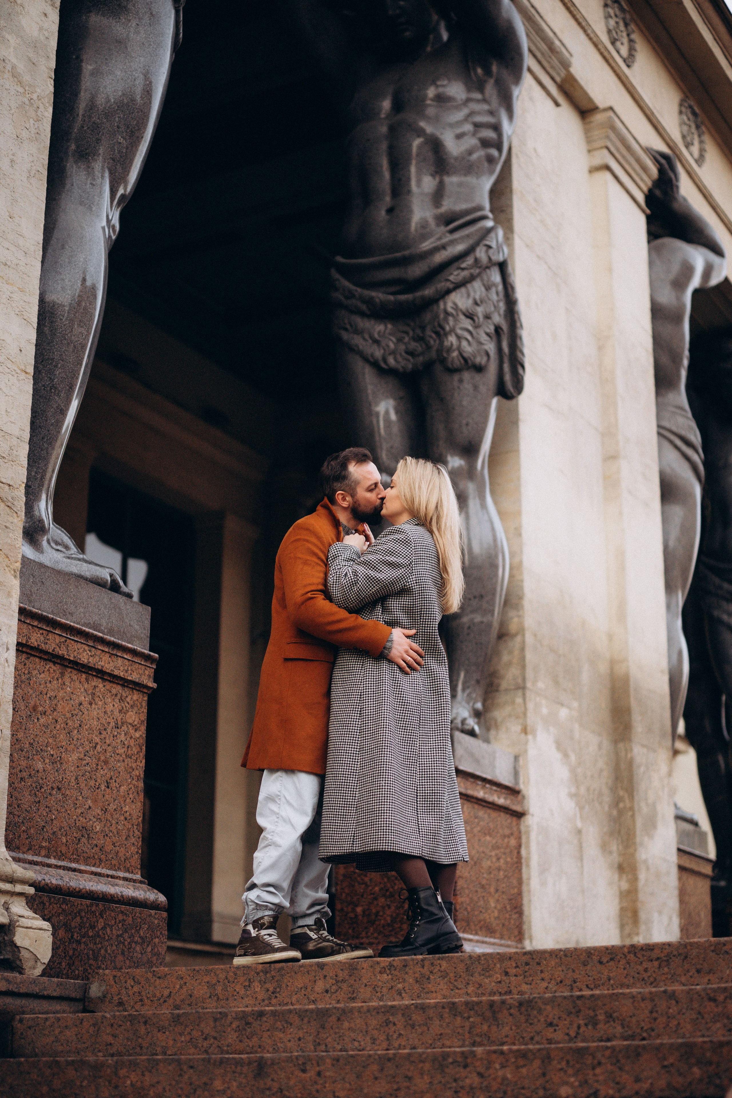 Лавстори Love Story в Санкт-Петербурге у Атлантов. Пара в коричневых пальто целуется на гранитной лестнице