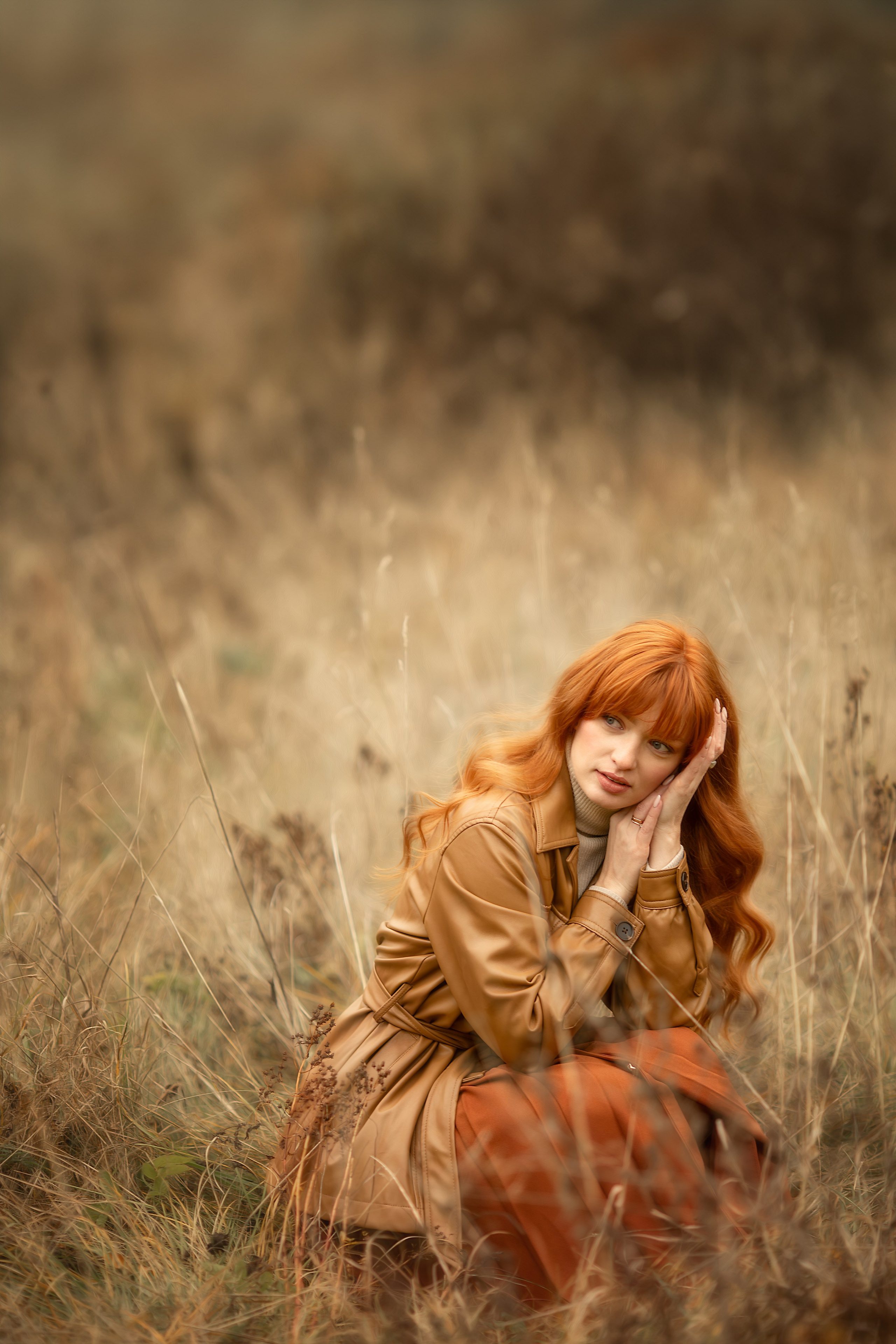 Девушка с рыжими волосами сидит в сухой траве и прикасается к лицу.