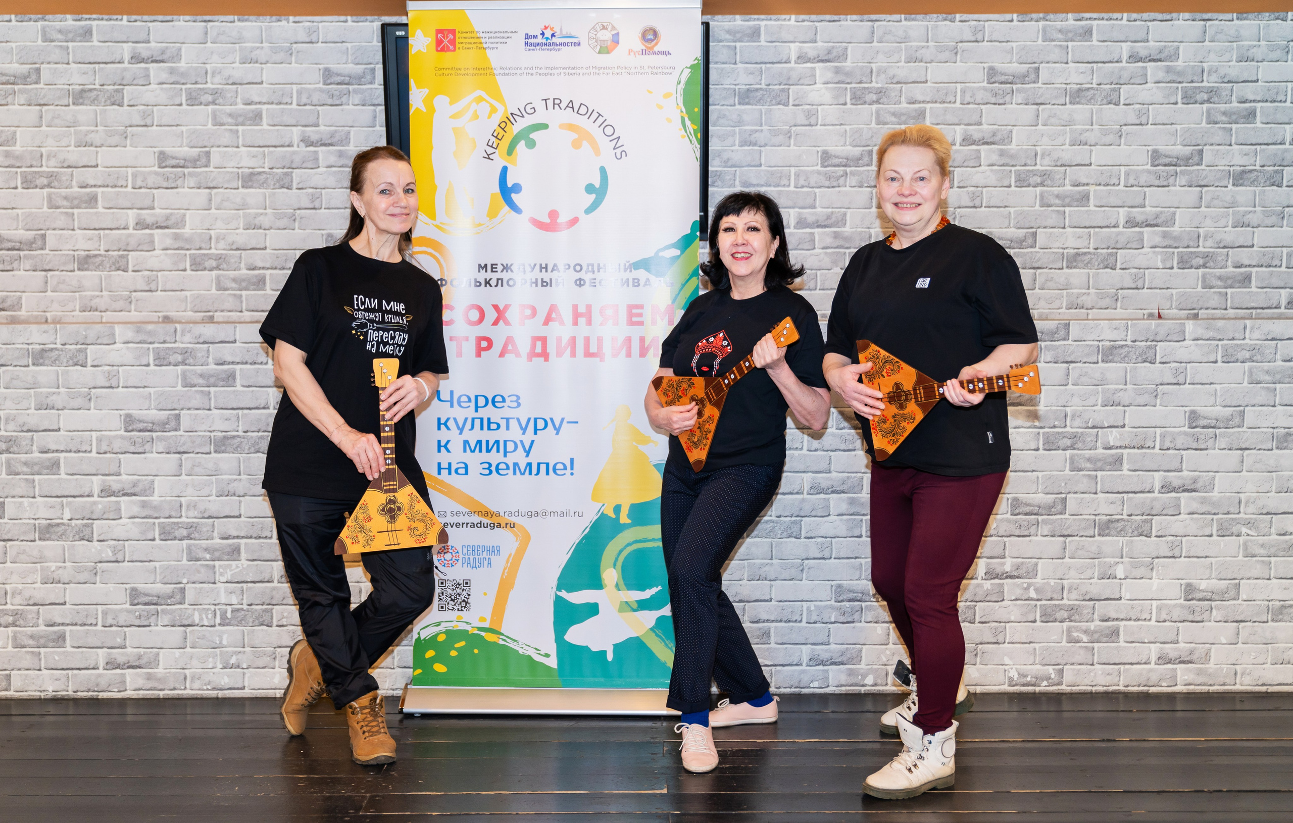 Три участницы XII Международного фольклорного фестиваля «Сохраняем традиции» Санкт-Петербург 2026 с балалайками на фоне баннера фестиваля. Фото: Мальцев Евгений, evrednix.ru.