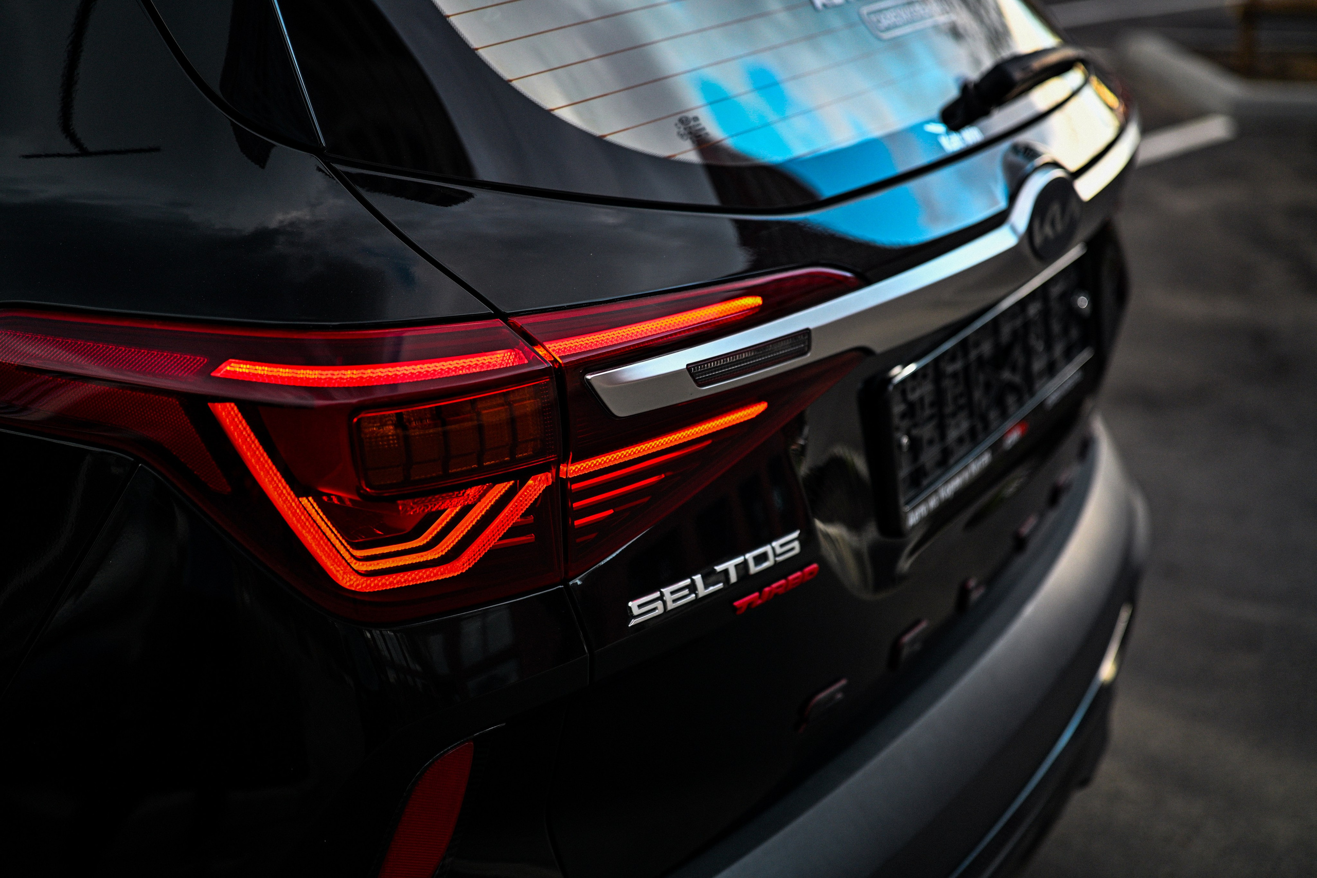Профессиональное фото автомобиля Kia Seltos в городских джунглях Новосибирска. Съемка с нижнего ракурса на фоне размытого делового центра. Акцент на агрессивный передний бампер и хромированные элементы кроссовера. Сине-оранжевая тонировка (цветокоррекция) создает атмосферу мегаполиса. Обзор экстерьера, автомобильный стайлинг, реклама корейского автопрома