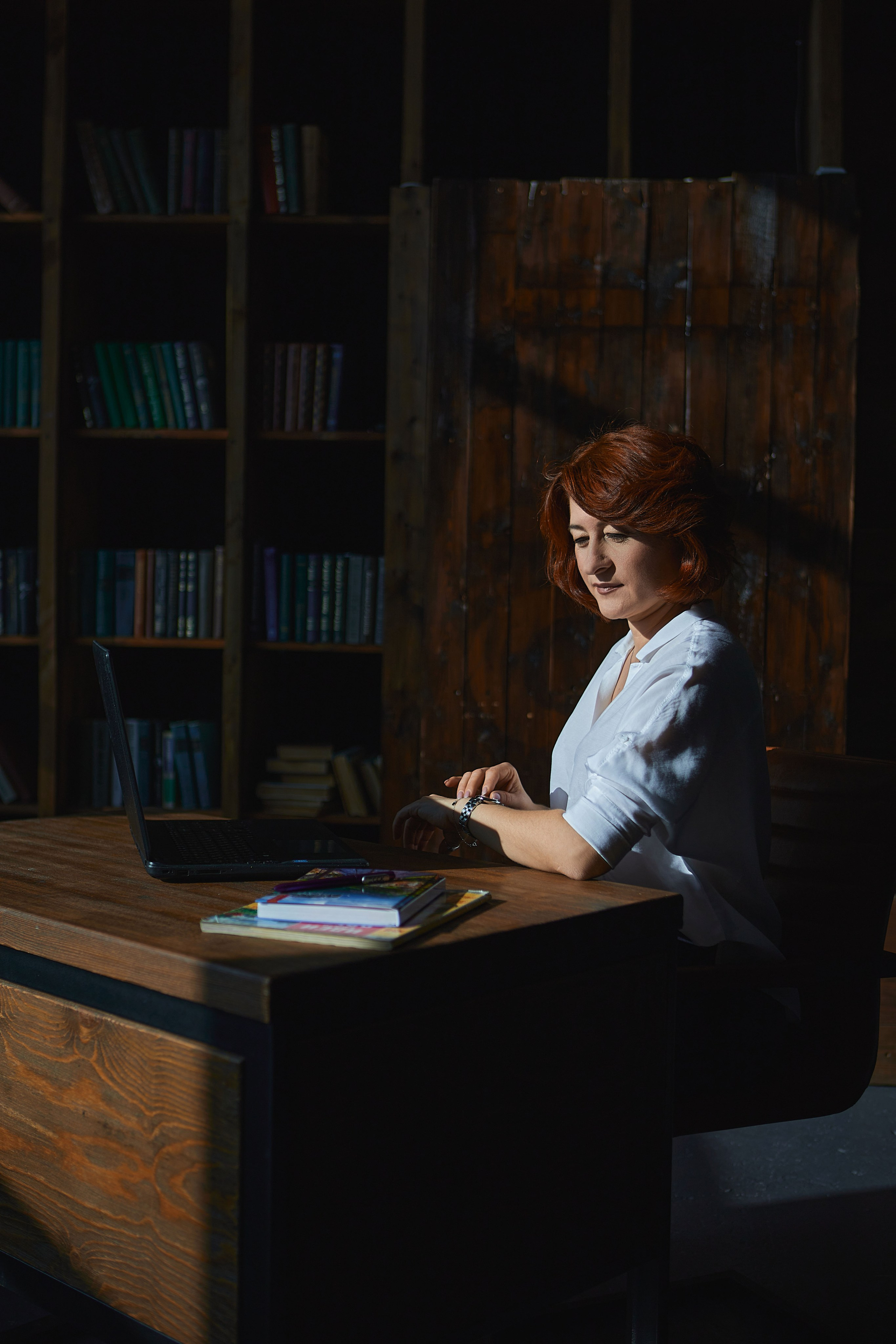 Женщина сидит за письменным столом на фоне книжного шкафа, на столе ноутбук