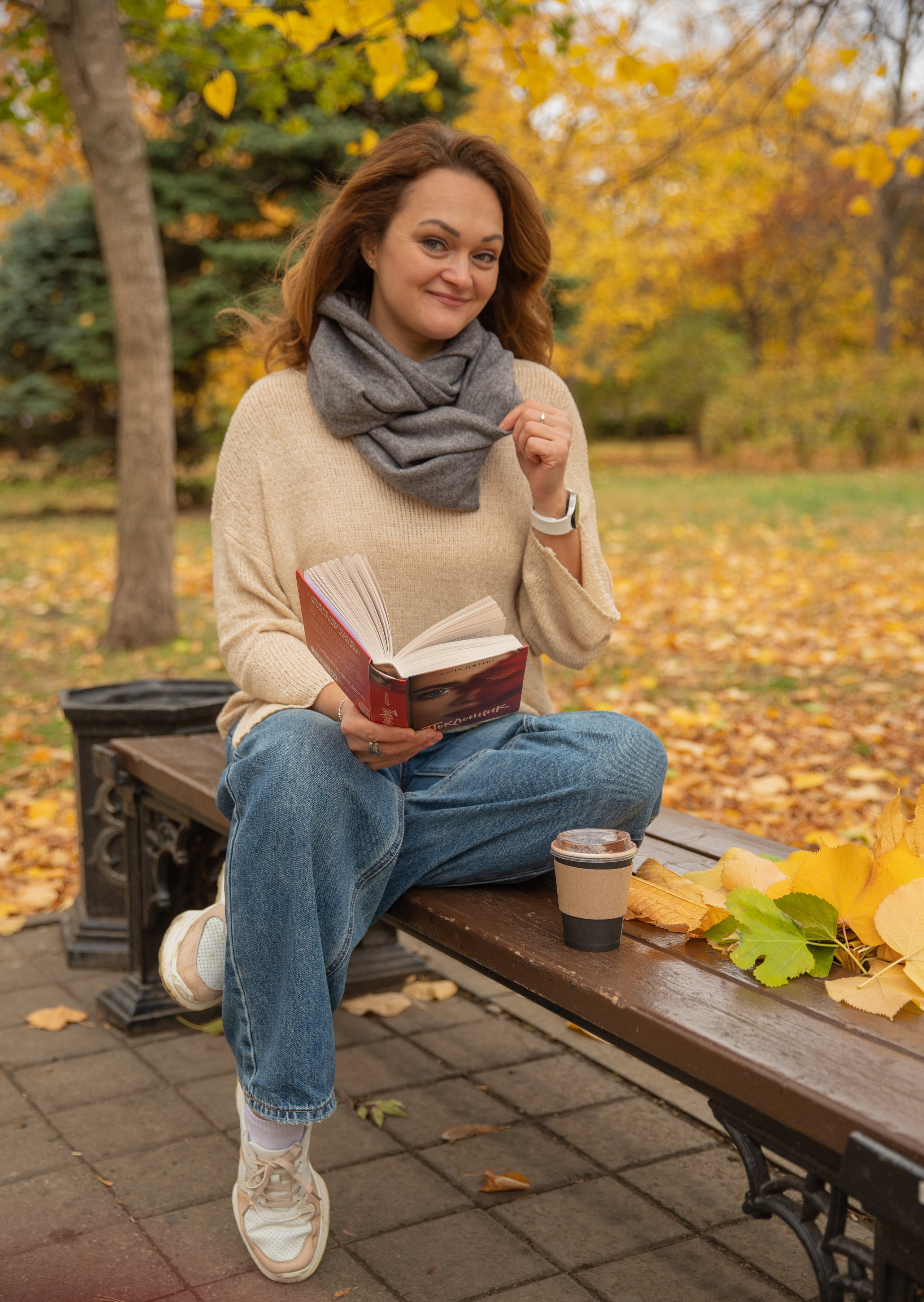 Стильная молодая женщина наслаждается чтением книги  во время осенней прогулки по аллеям парка Чистяковская роща, усыпанным золотыми листьями.