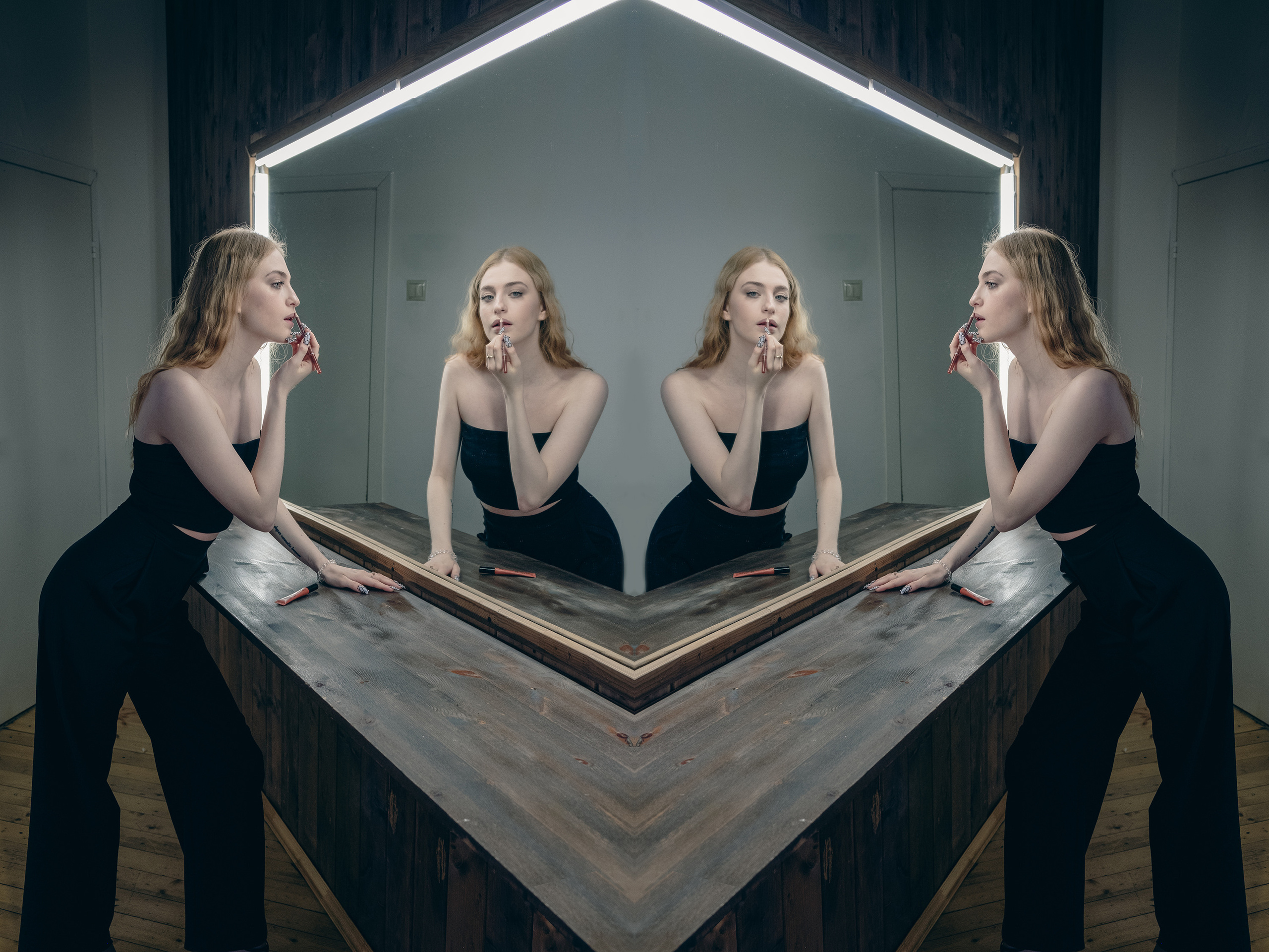 Девушка,блондинка,в черном топе и черных брюках стоит у зеркала в студии и подкрашивает губы..Фото отзеркалено по центру..Необычный эффект..