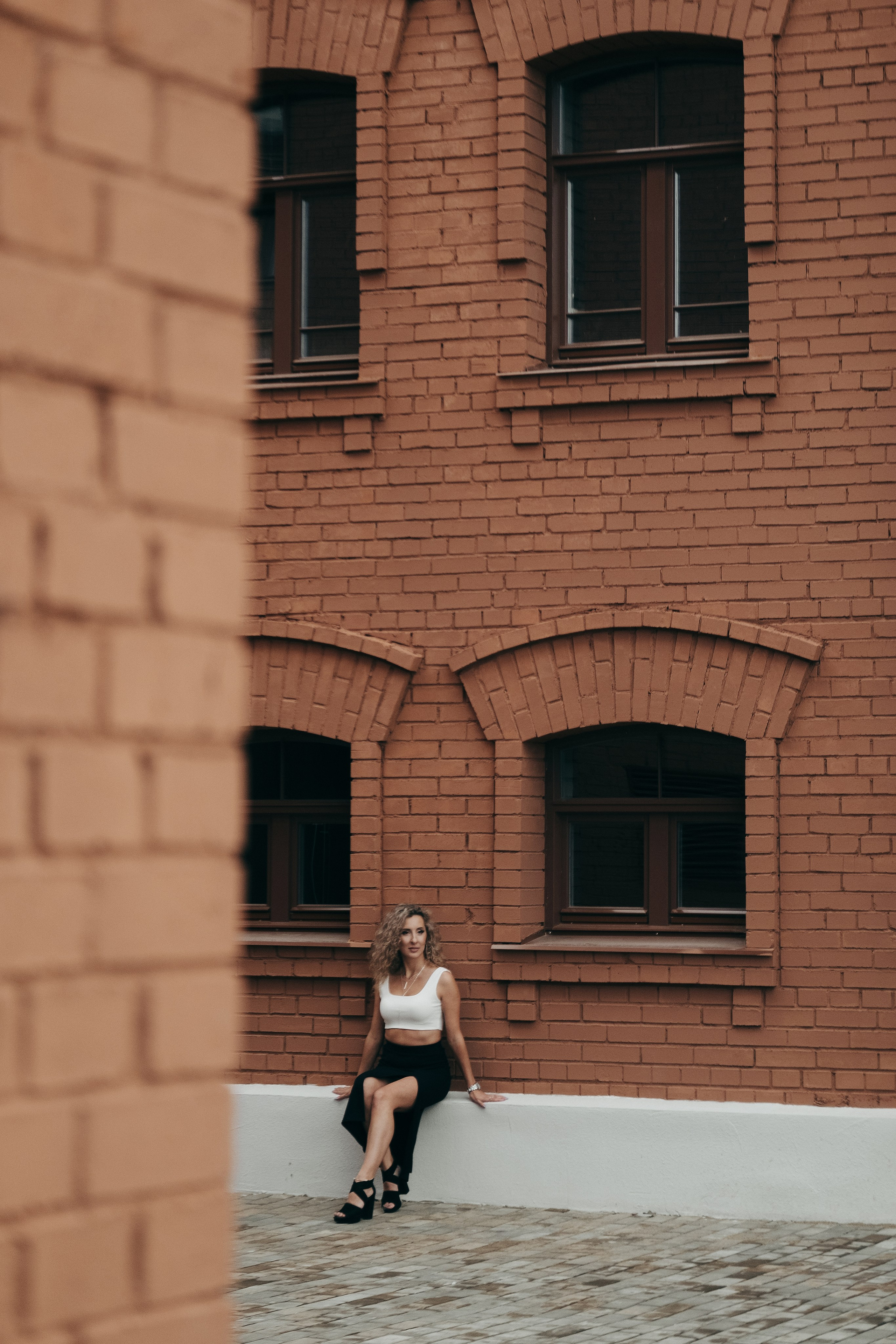 Фотограф в Краснодаре и Архангельске Денис Ожигин. Индивидуальная фотосессия для кудрявой девушки блондинки в городских локациях на фоне кирпичного здания из красного кирпича в белом платье
