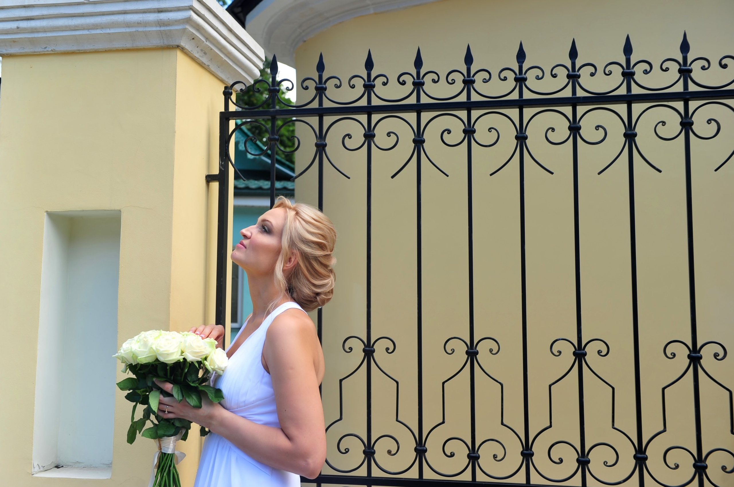 Свадьба на Воробьёвых Горах рядом с МГУ, невеста стоит с букетом кремовых роз у кованной ограды и желтой стены храма