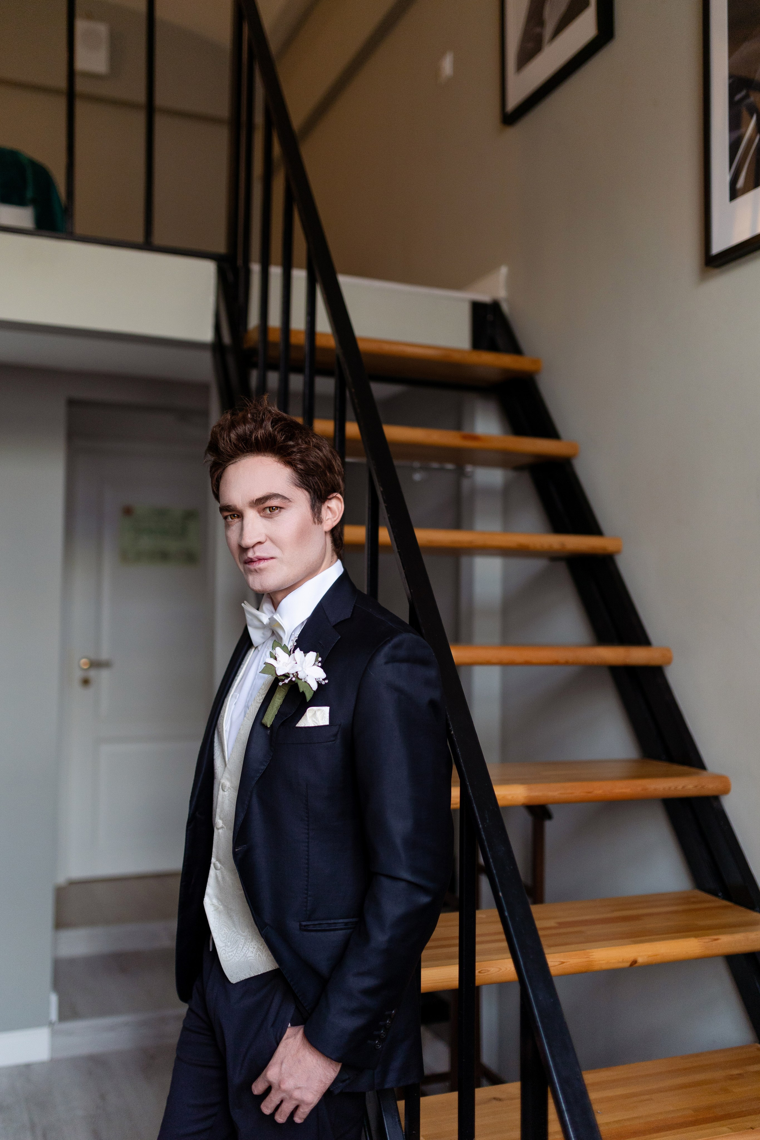 Жених стоит у лестницы в белоснежной рубашке и черном костюме.