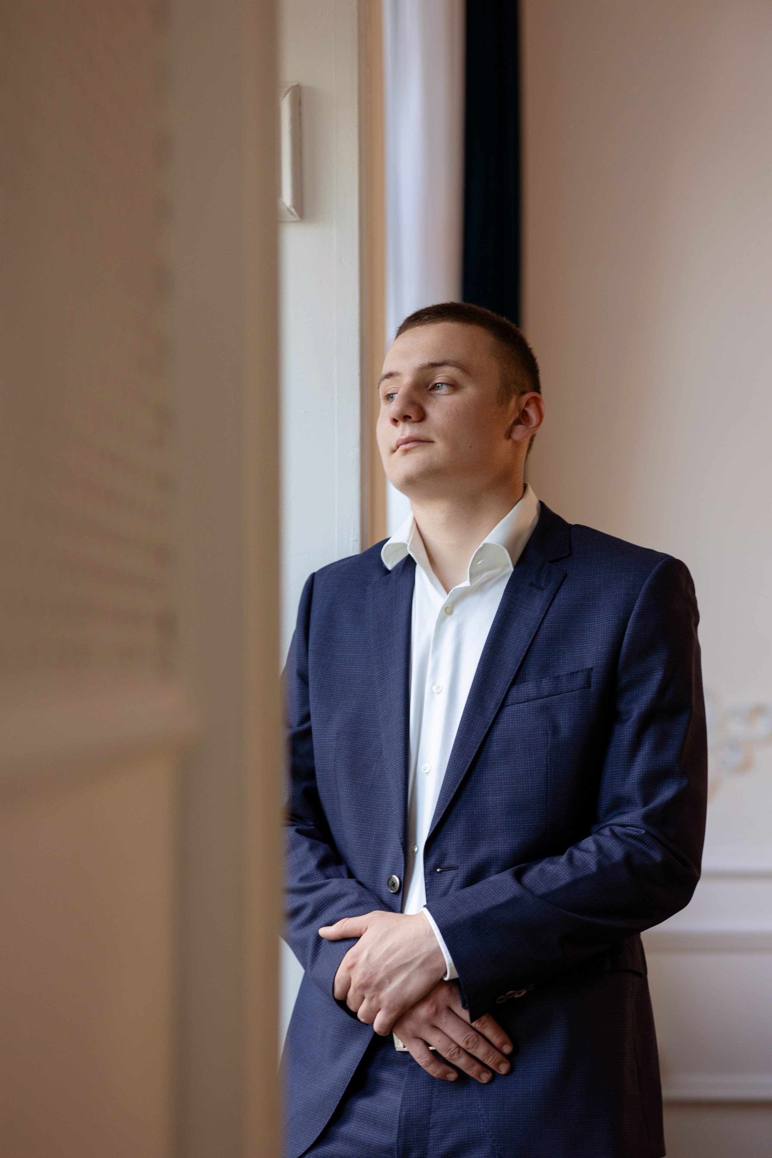 Жених в строгом черном костюме стоит у окна.