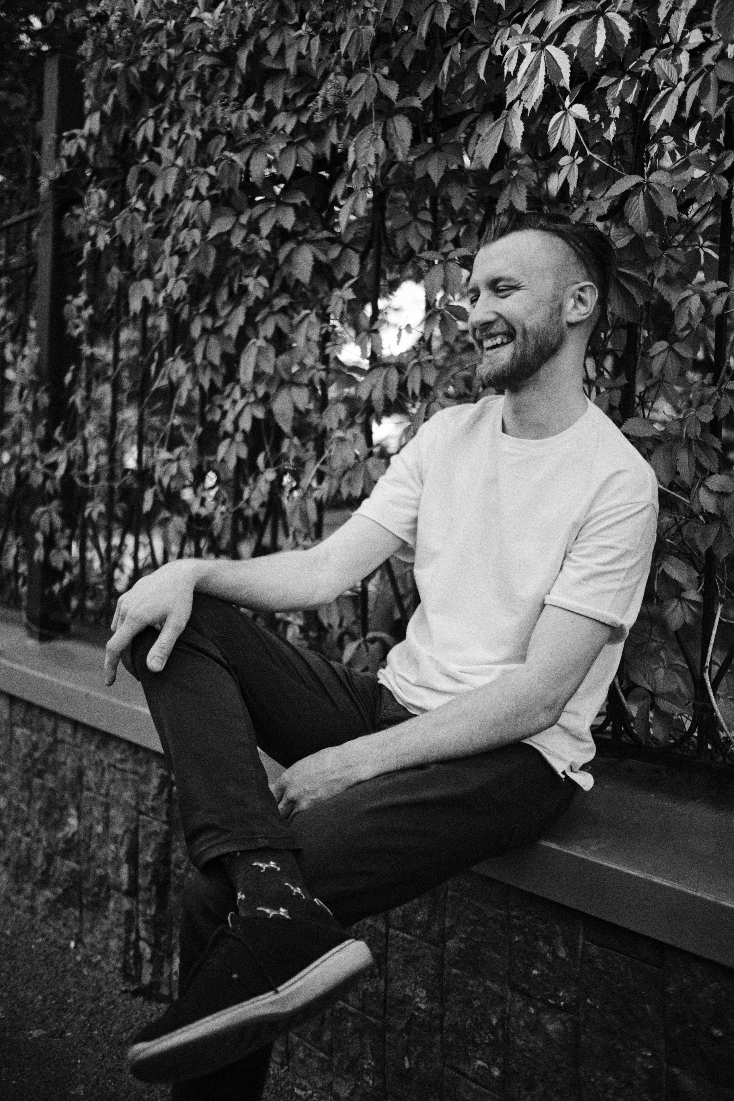 Мужчина смеется, сидя на парапете забора, оплетенного диким виноградом