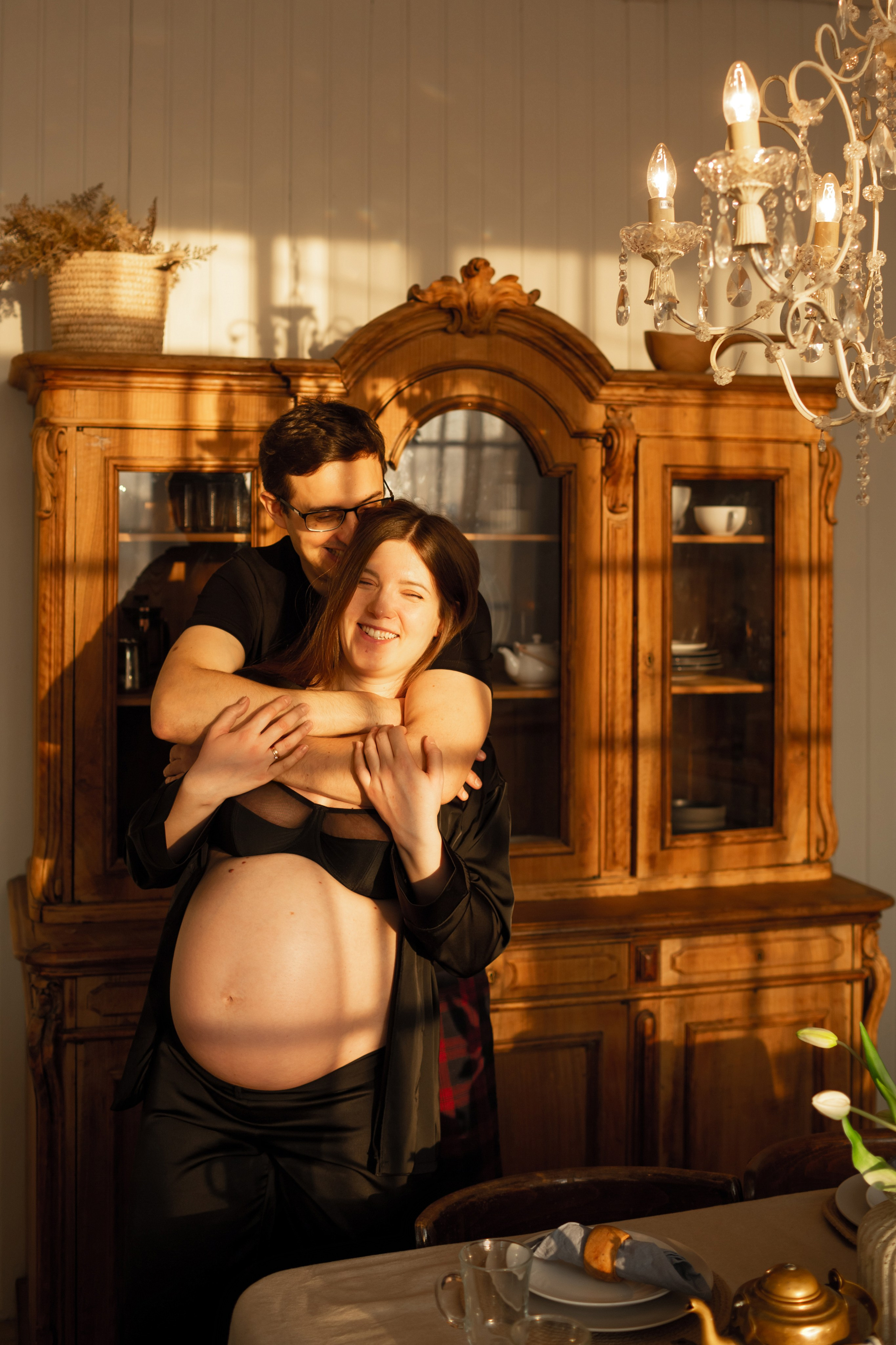 Мужчина обнимает беременную женщину в солнечном свете у деревянного серванта