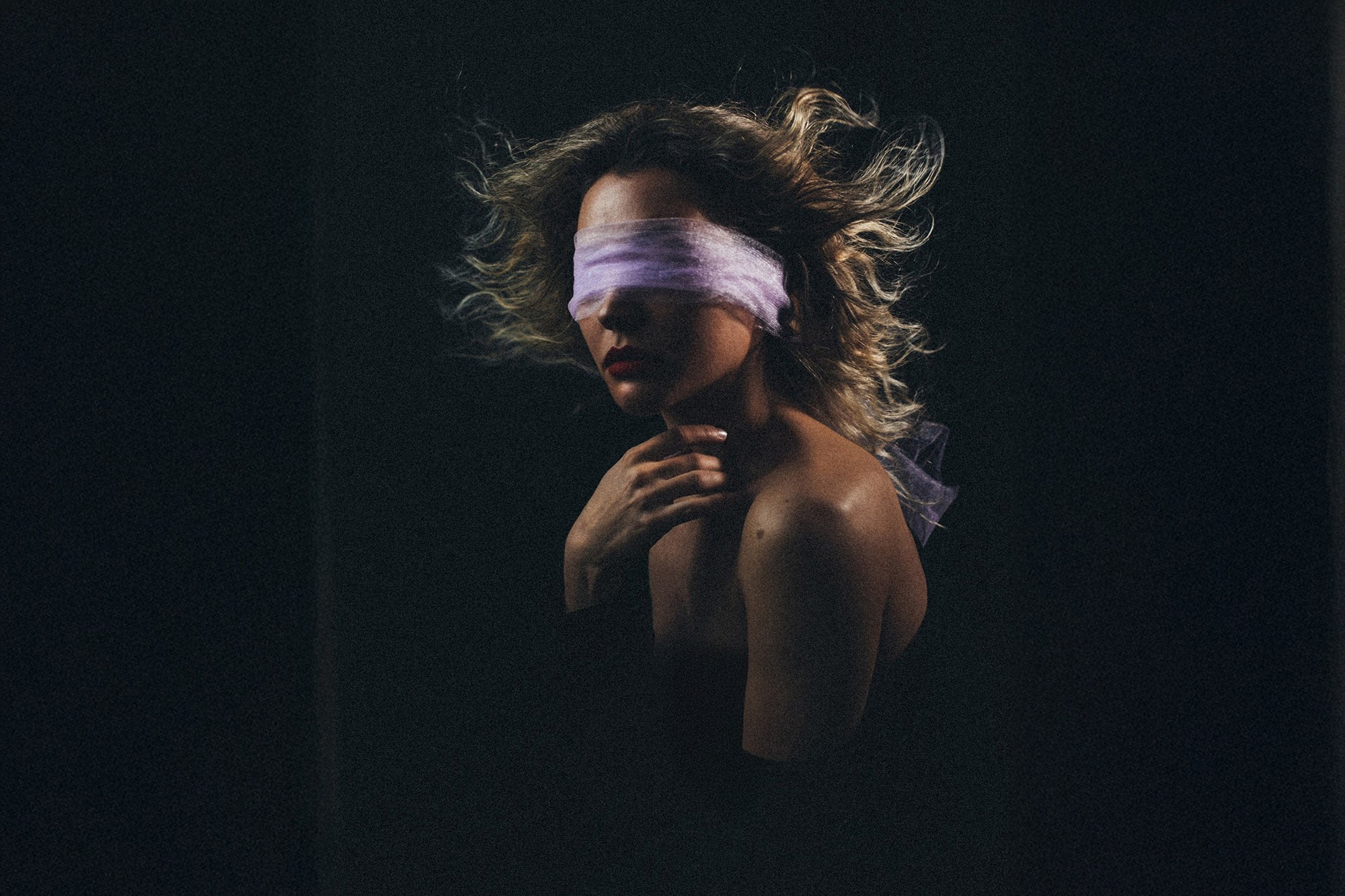 Художественный портрет женщины в профиль на темном фоне. Глаза закрыты полупрозрачной сиреневой повязкой, волосы развеваются, создавая эффект динамики. Драматичное освещение подчеркивает линию плеч и задумчивый образ. Фотограф: Галина Скорик, Сочи.