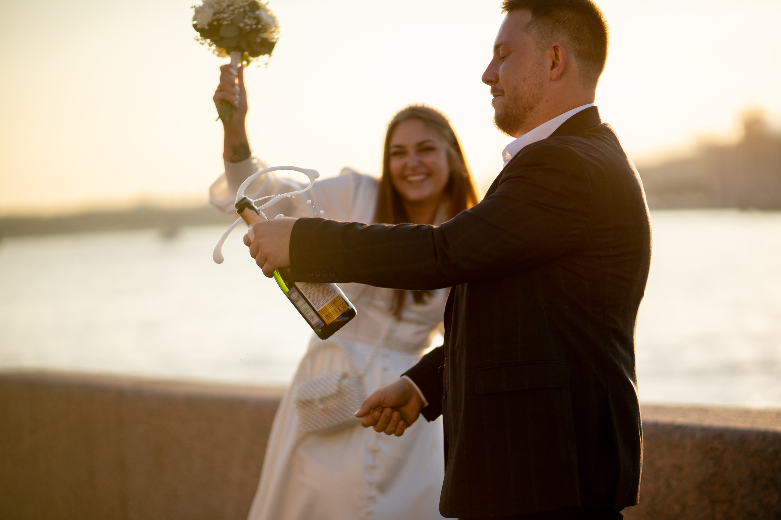 невеста поднимает руку с букетом вверх жених открывает шампанское на набережной невы на закате