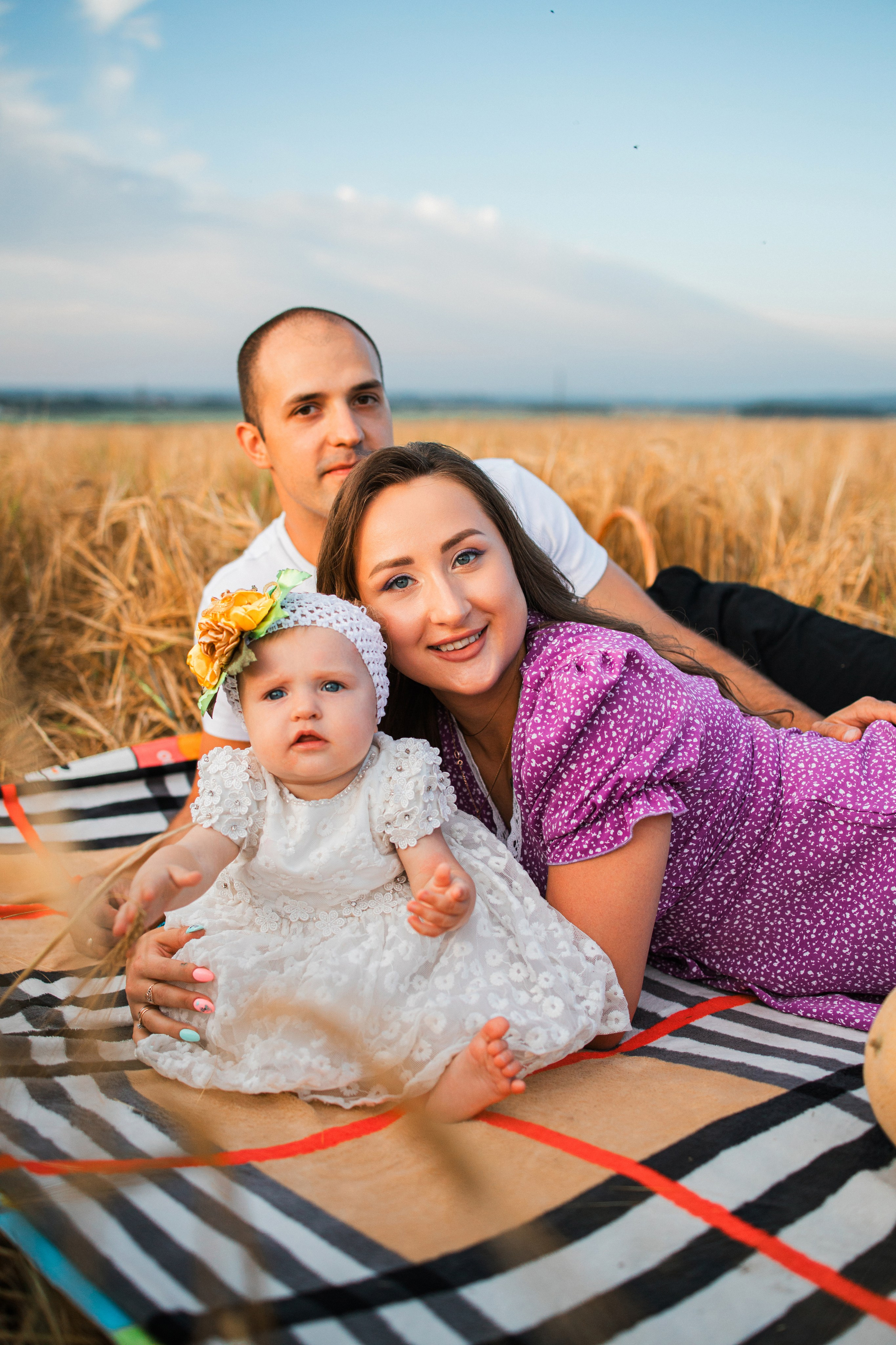 Папа и мама обнимаются, дочка ползает рядом на пледе в поле при закате.