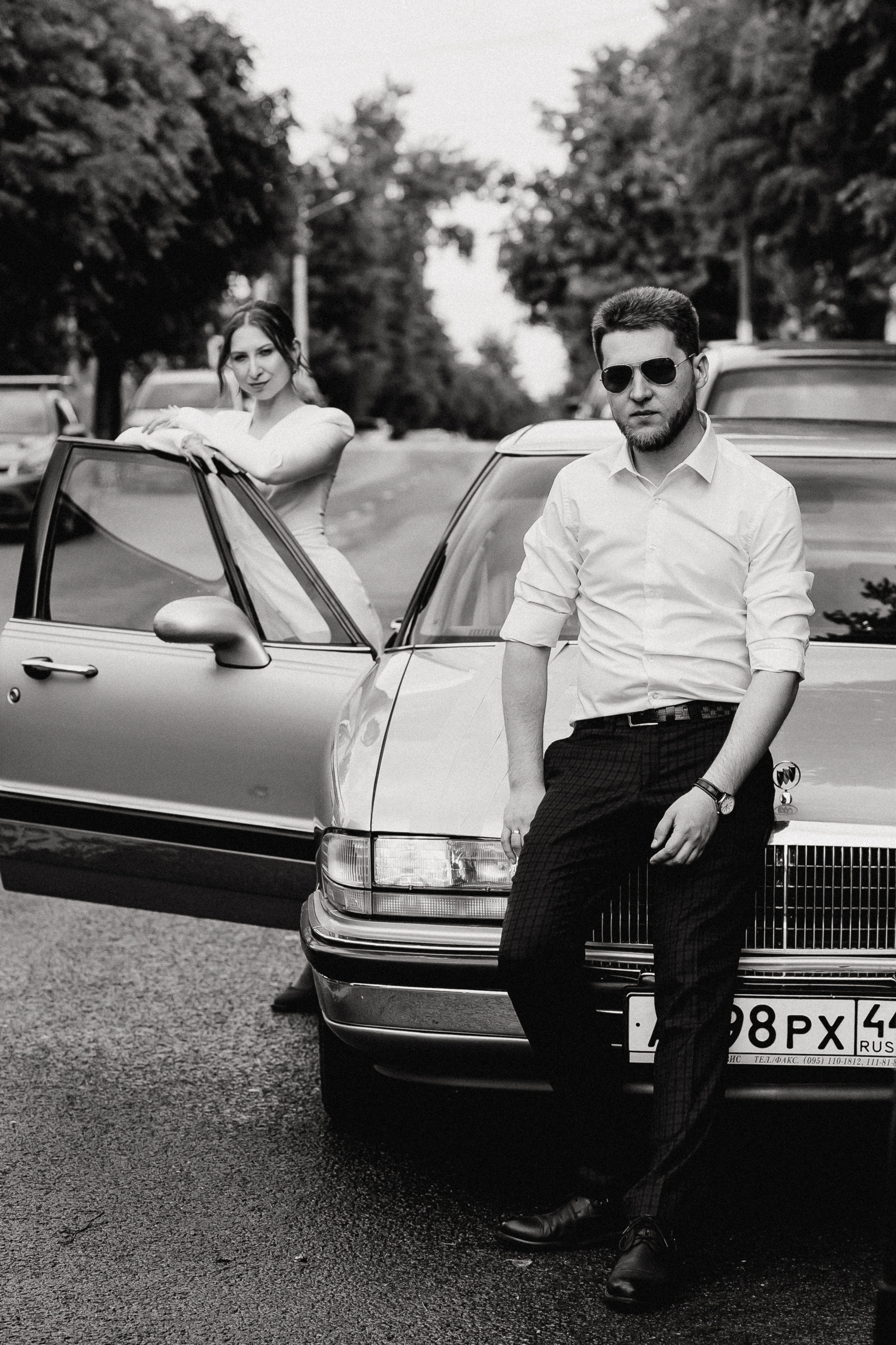 Мужчина сидит на капоте свадебного автомобиля, девушка опирается на дверь автомобиля 