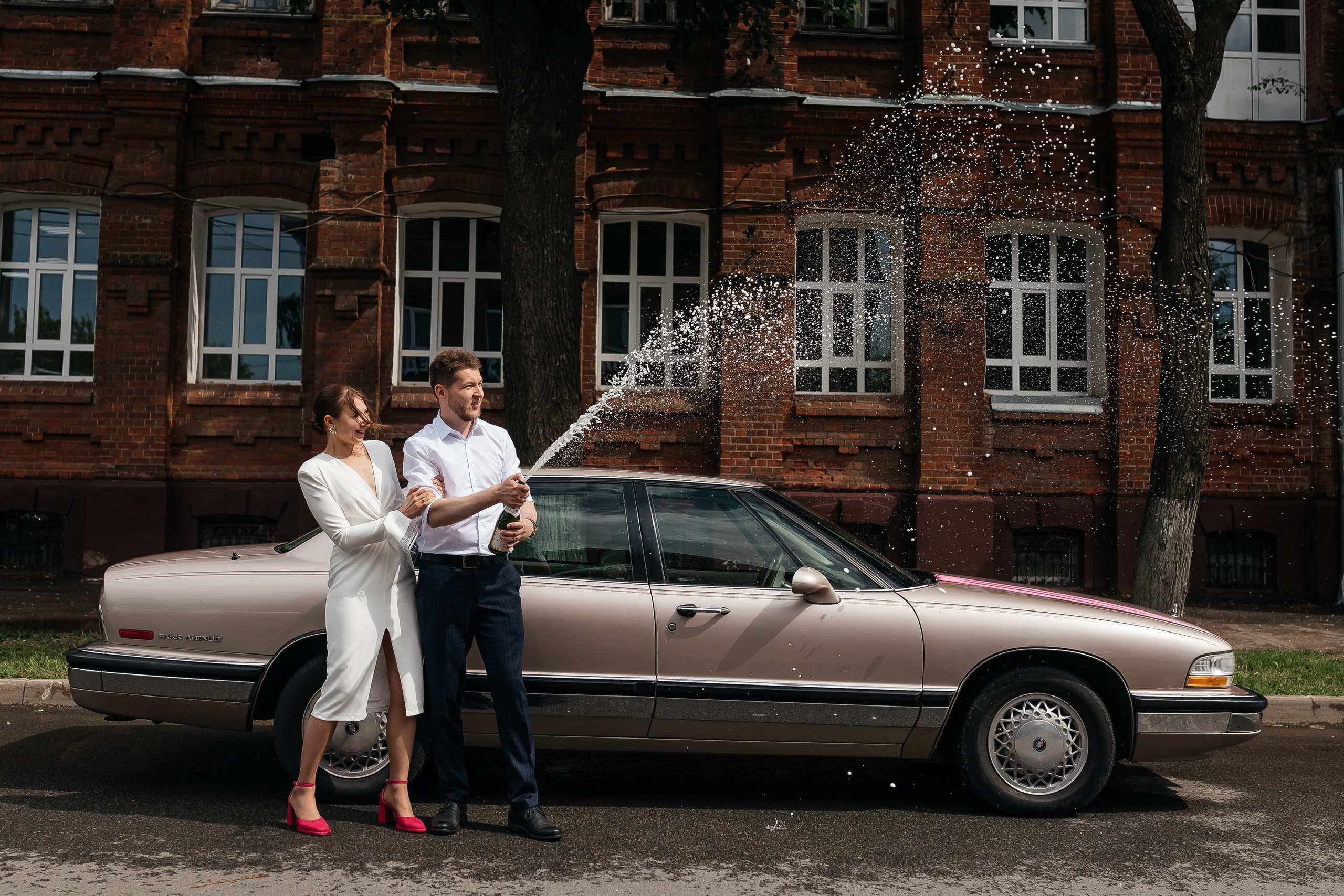 Жених и невеста на фоне свадебного автомобиля открывают шампанское