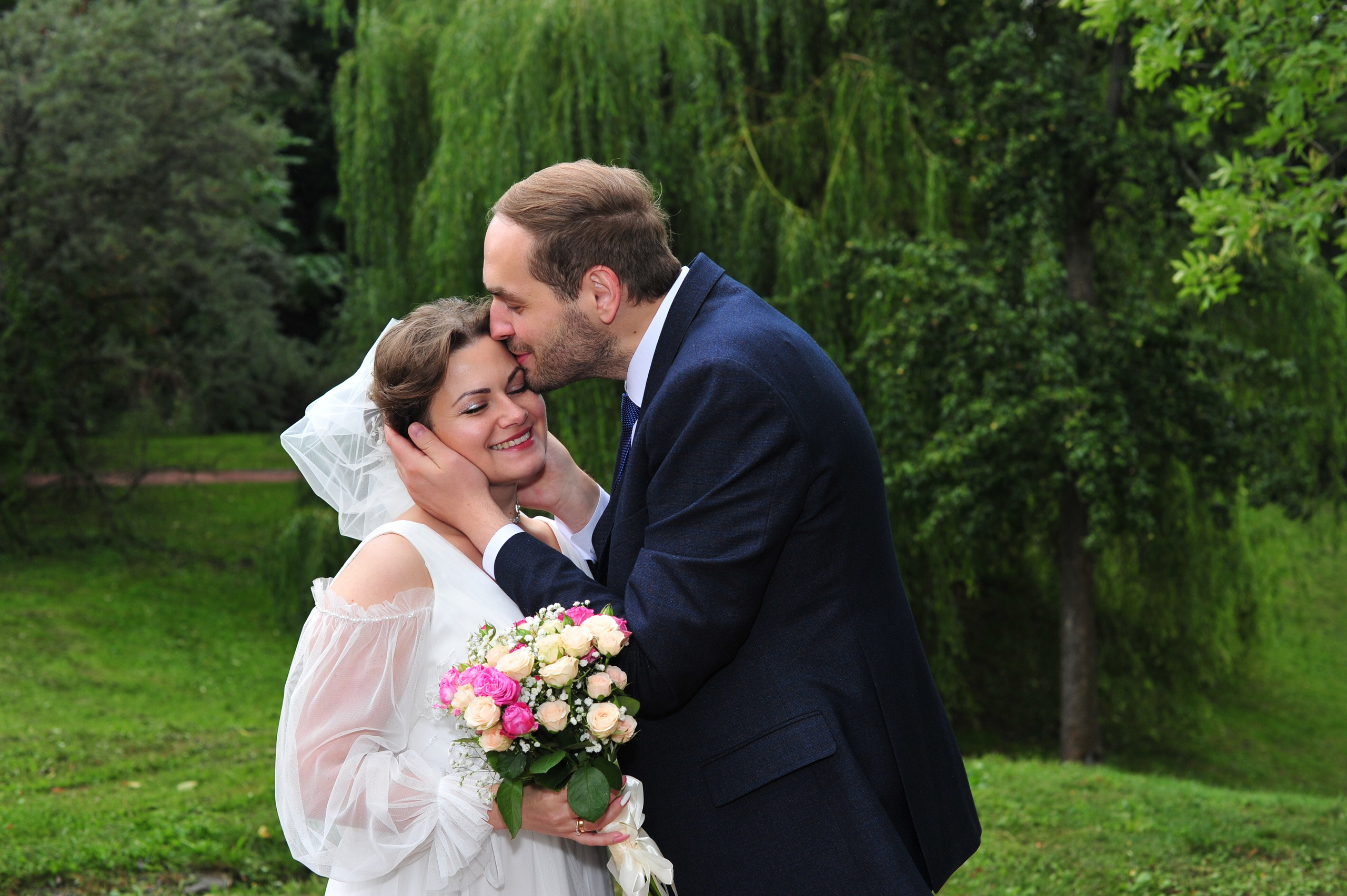 Свадьба в Царицыно пасмурным летним днем, жених нежно целует невесту в лоб на фоне изумрудной зелени 