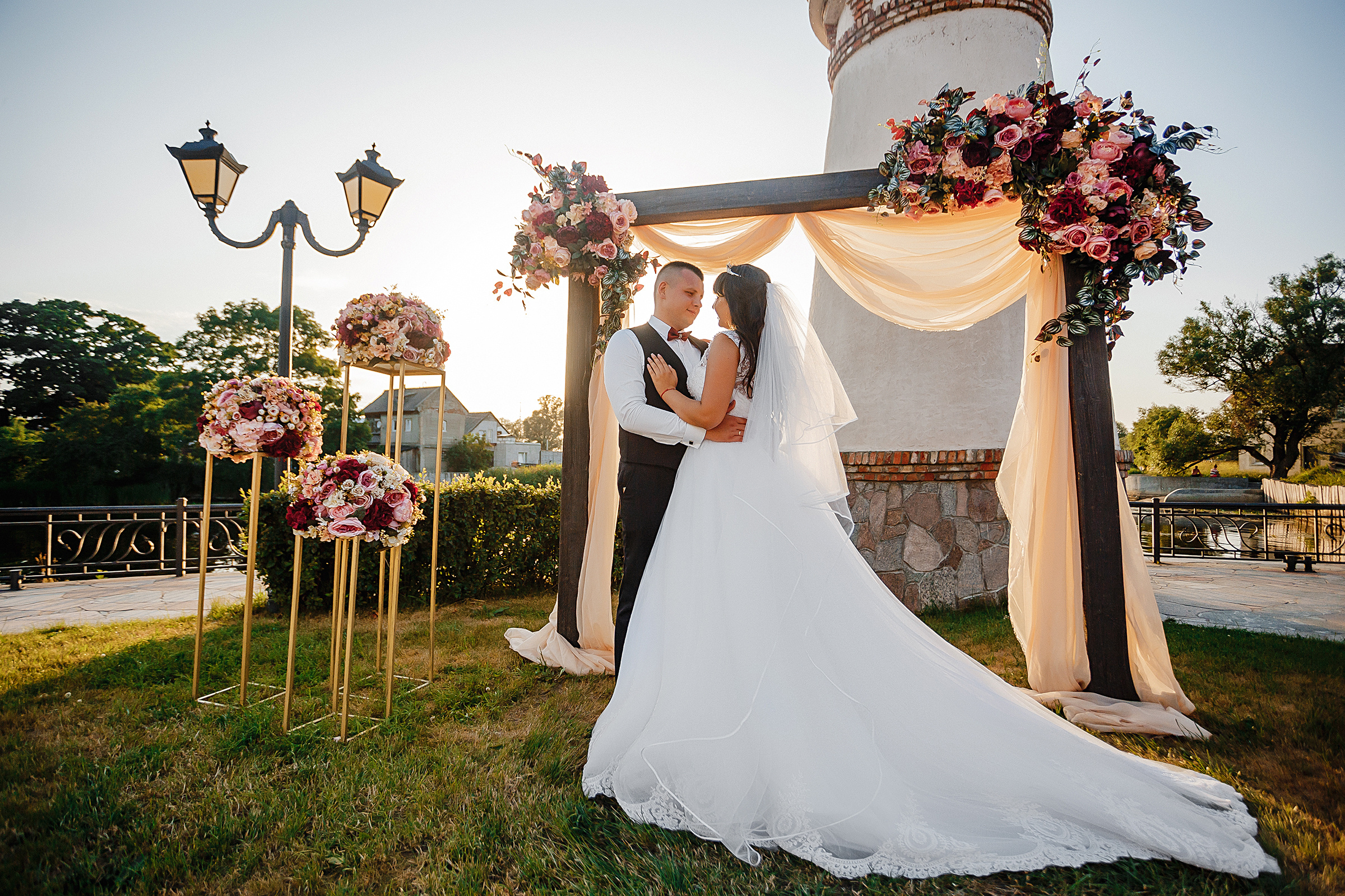 Молодожены стоят у арки выездной регистрации на закате