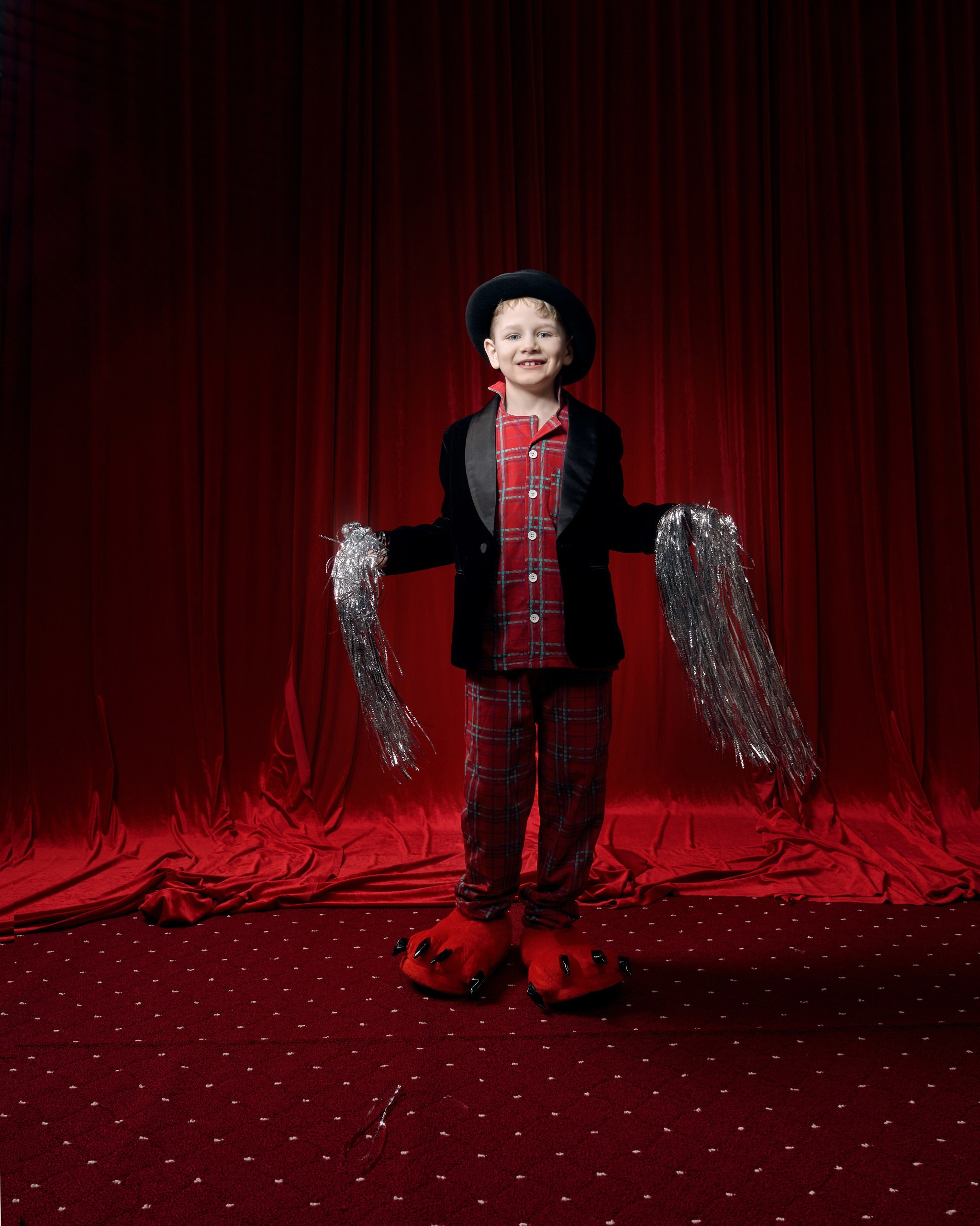 Детская фотосессия: мальчик в костюме на фоне театрального занавеса. Профессиональная студийная съемка для детей: веселый мальчик в необычном образе — пижама, пиджак, шляпа и лапы-тапочки — позирует на красной сцене. Идея для семейного фотоальбома от семейного фотографа. Фотосессия для детей, полная сюрпризов: мальчик в пижаме и шляпе на красной сцене. Семейный фотограф помогает раскрыть детскую фантазию и создать уникальные кадры.